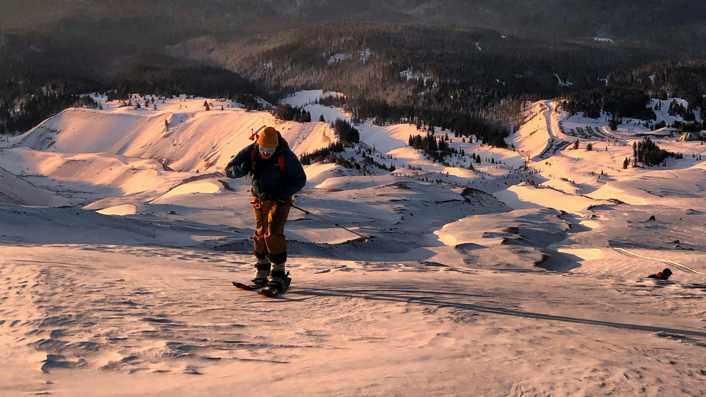 A snowboarder walking up a hill at sunrise.
