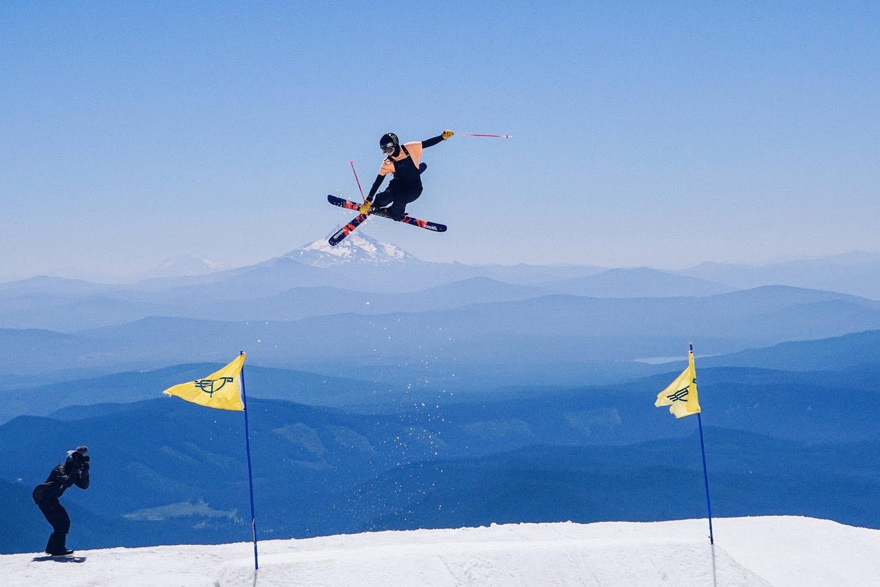 Maggie jumps and crosses her skis in their air, bringing her legs up to her chest. A tall mountain peak is visible in the background. 
