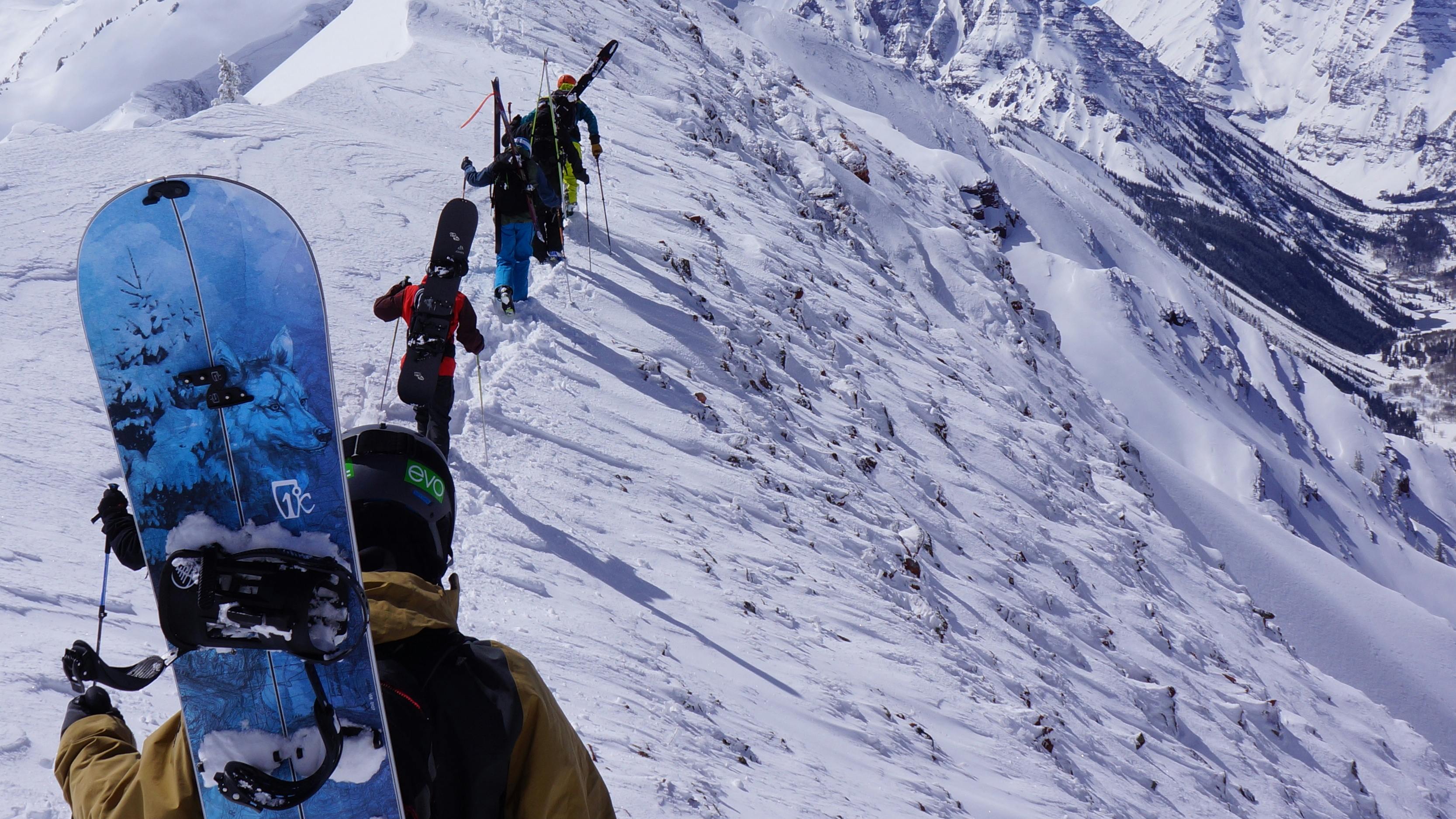 A line of snowboarders traverse a ridge with their snowboards on their backs