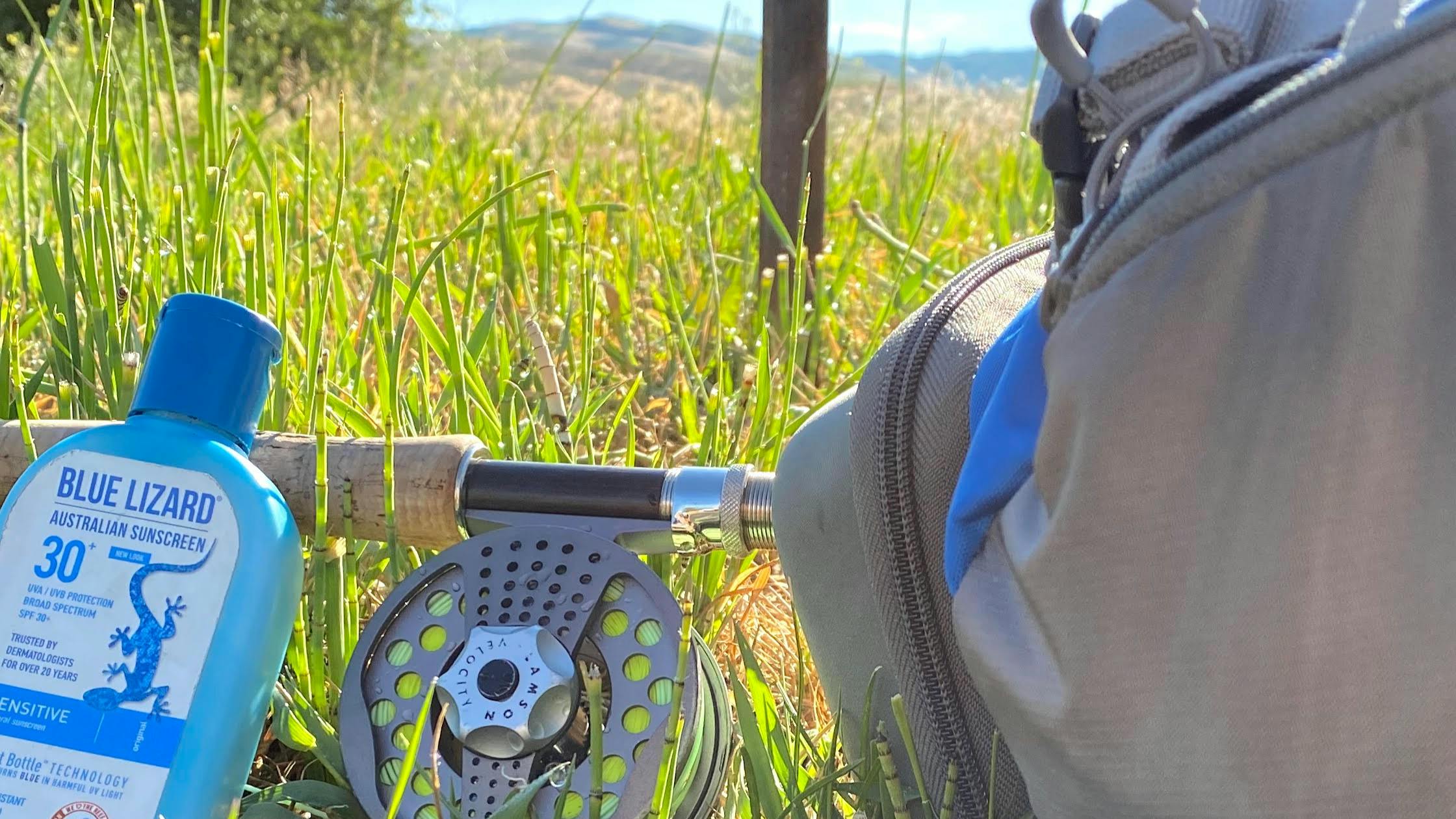 A fly fishing pack, sunscreen, and rod and reel laying in the grass