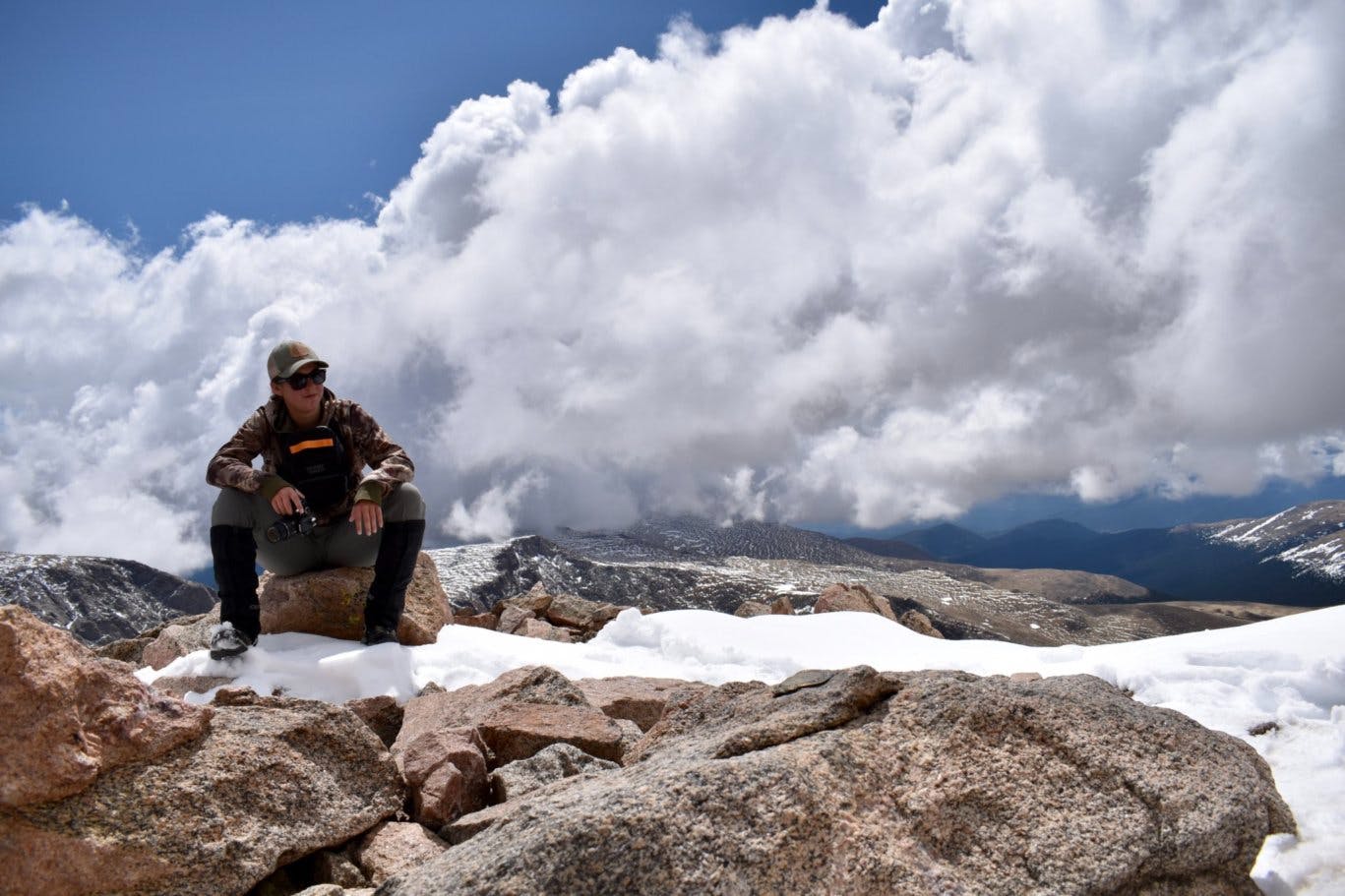 A hiker sitting on a rock on a snowy mountain summit with white puffy clouds in the background