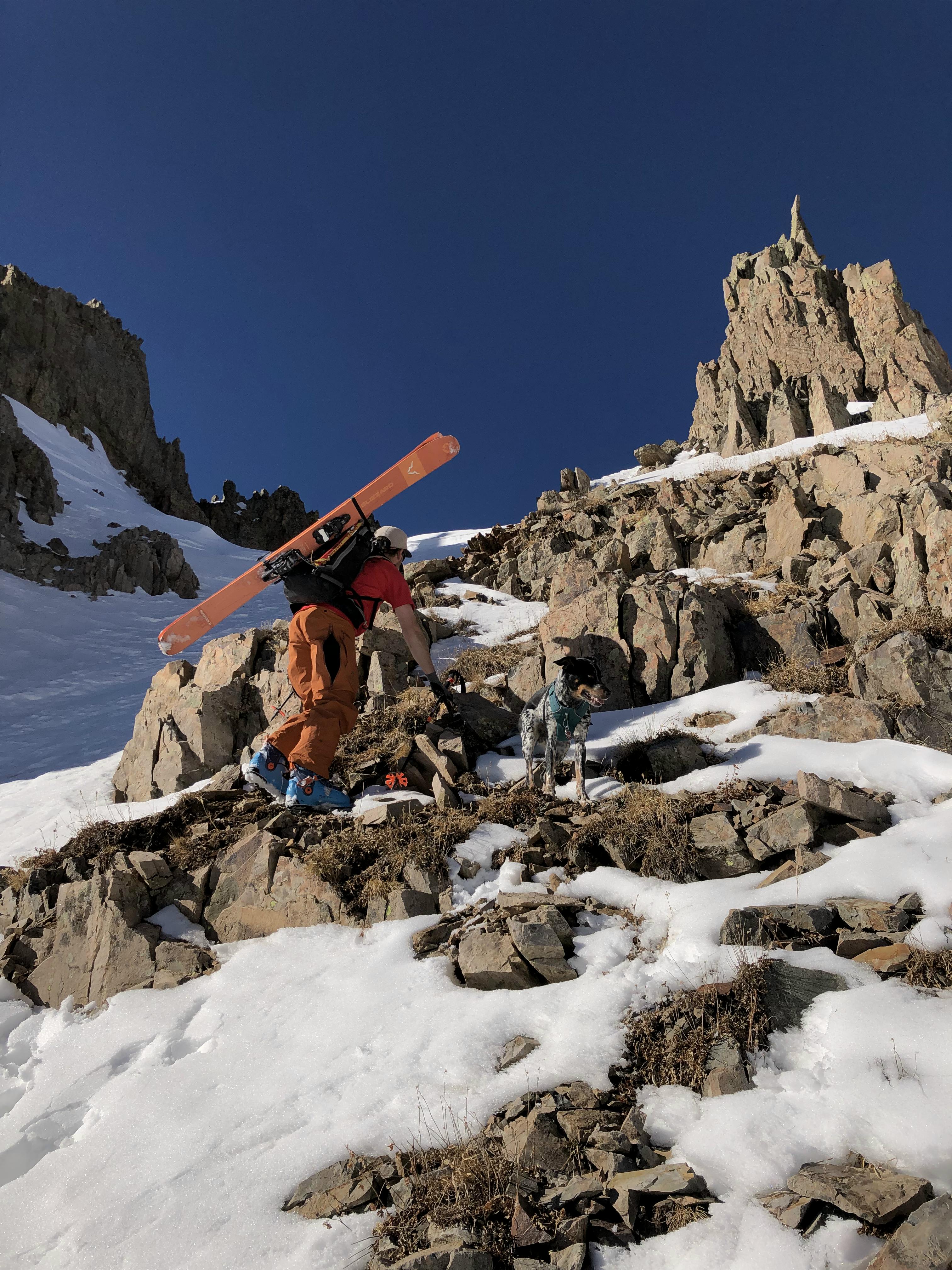 A skier climbing up a mountain with the Blizzard Rustler 11 Skis · 2022 skis on his back.