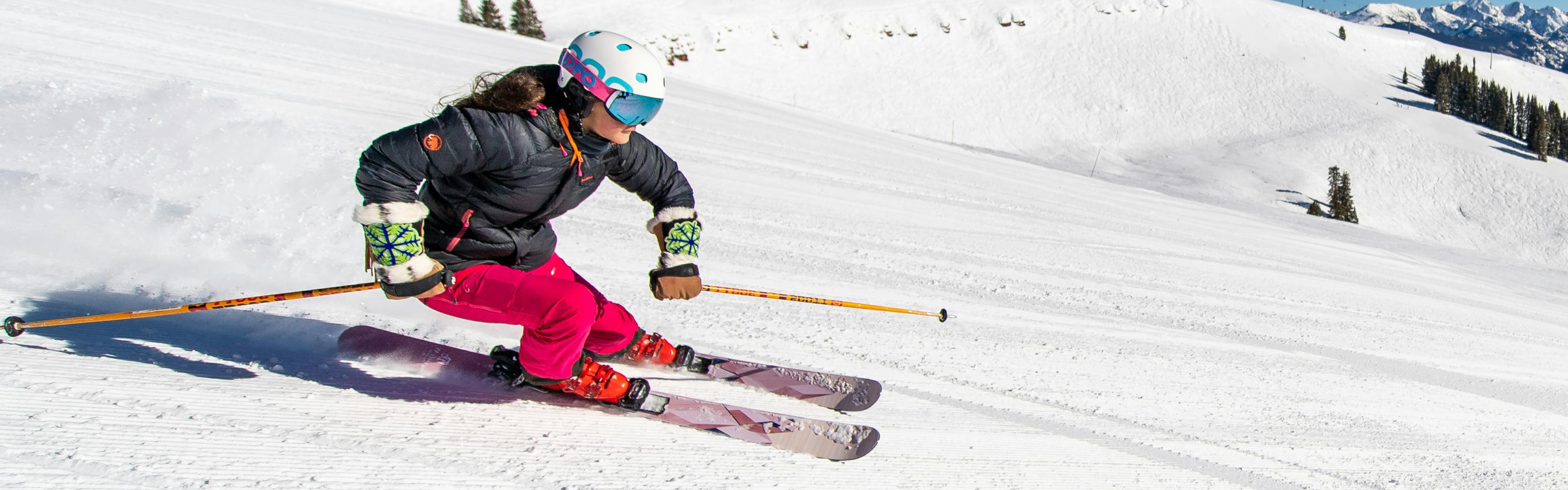A woman in hot pink ski pants and a black jacket on purple skis and with orange poles skiing down a groomer slope.