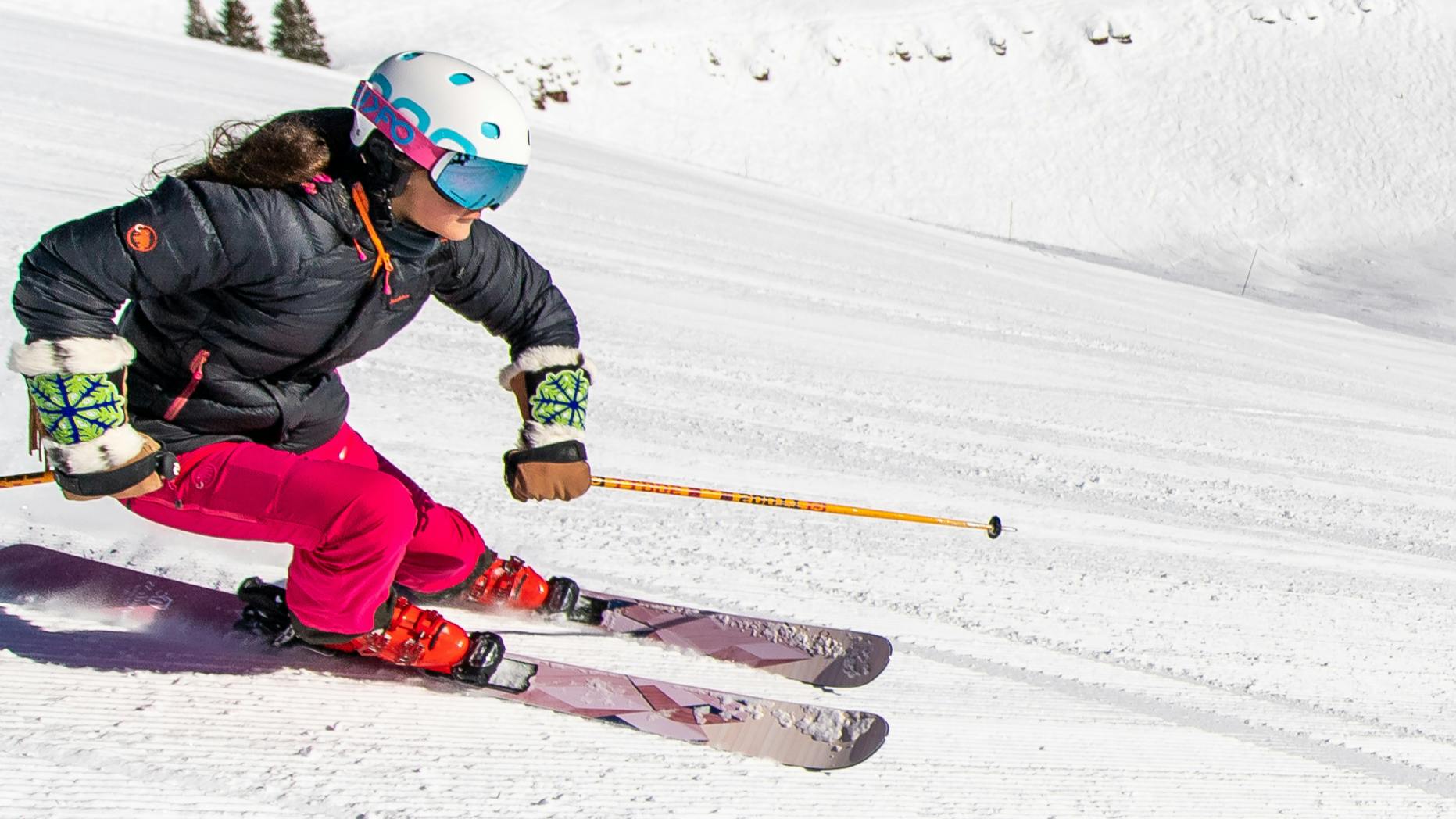 A woman in hot pink ski pants and a black jacket on purple skis and with orange poles skiing down a groomer slope.