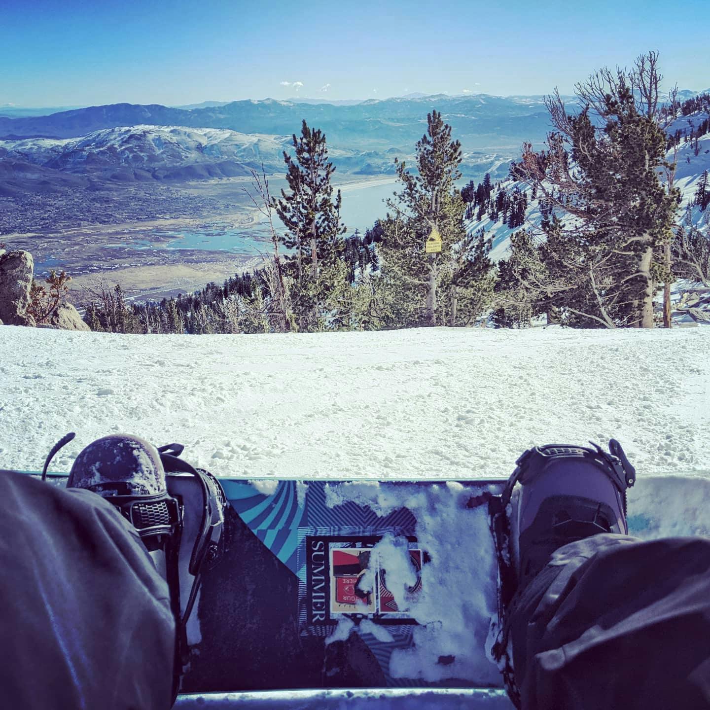 Top down view of a snowboard at the top of a ski run. You can see the valley and mountains in the background. 