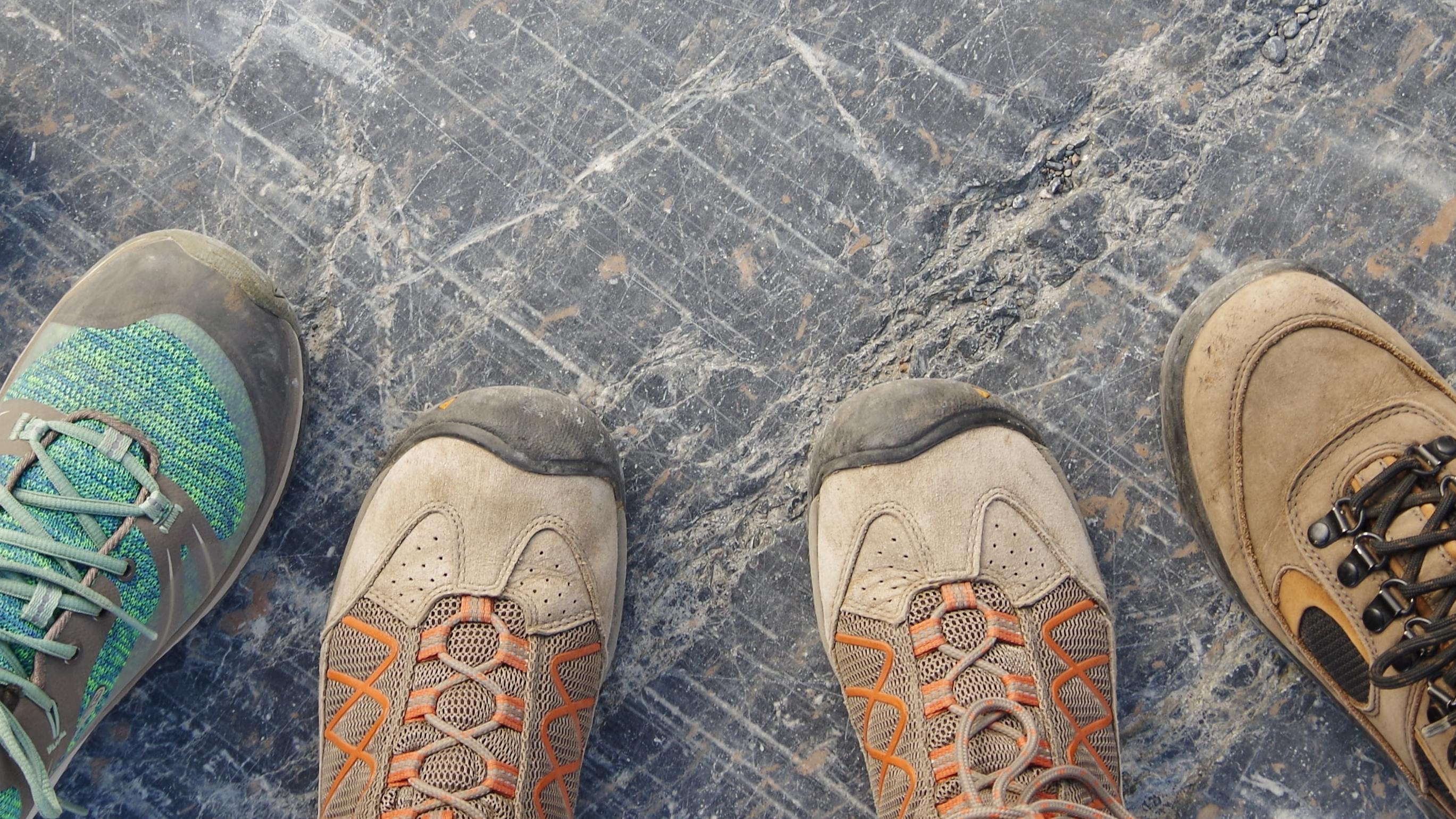 Three people stand next to each other on rock that's been scraped by a glacier. We look down at their shoes which are arranged in a semi-circle. They're wearing trail runners and hiking boots.