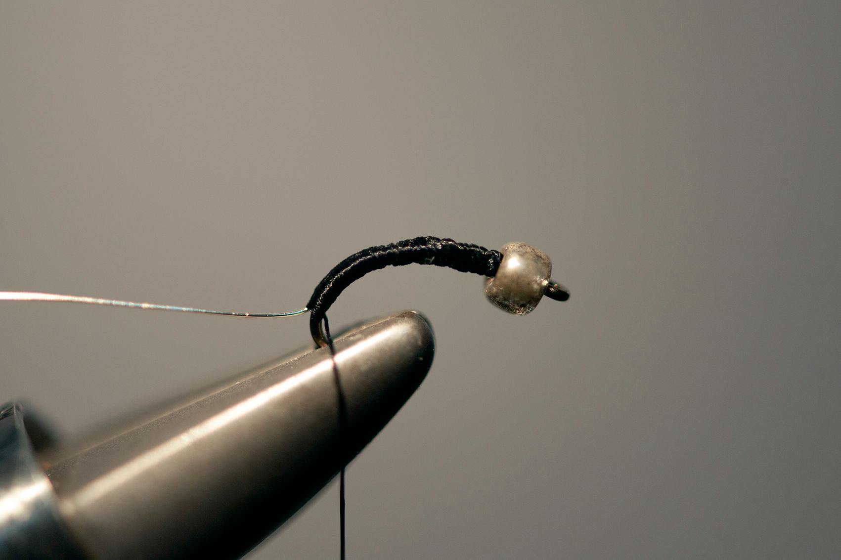 The same hook and bead and thread from above is pictured in the vice jaws but now there is even more thread and the fly looks almost complete. 