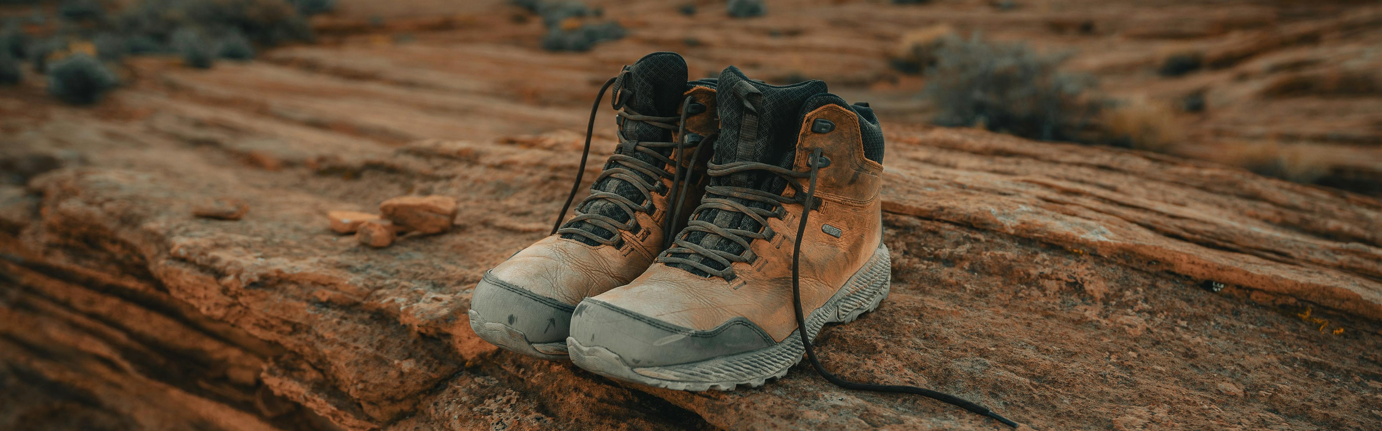A pair of dusty brown boots sits on a rusty-red rock formation. The laces are untied.