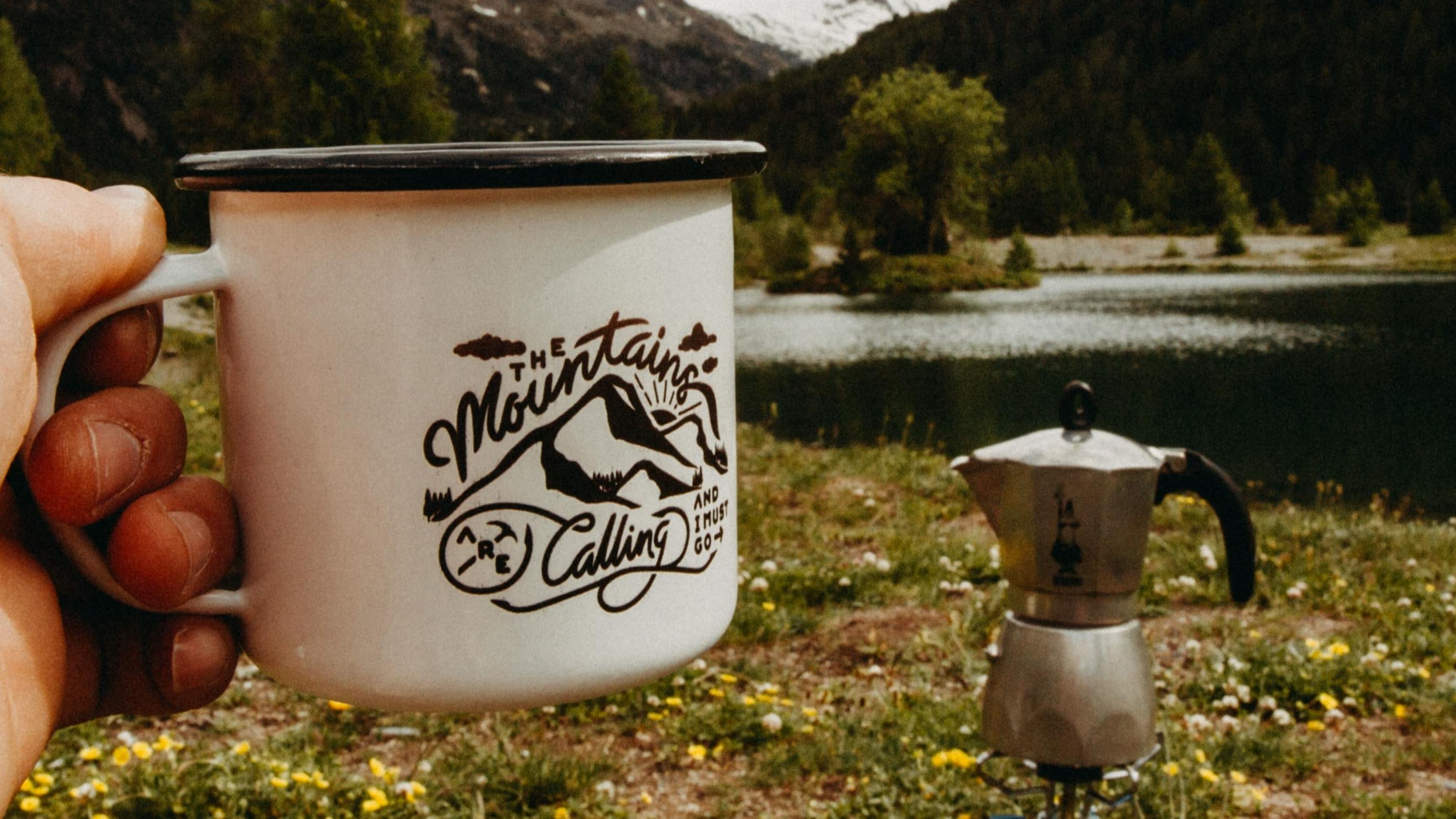 Someone holds out a tin mug that reads "The Mountains Are Calling and I Must Go." In the background, a Moka pot coffeemaker sits on a meadow floor with a river behind it. 