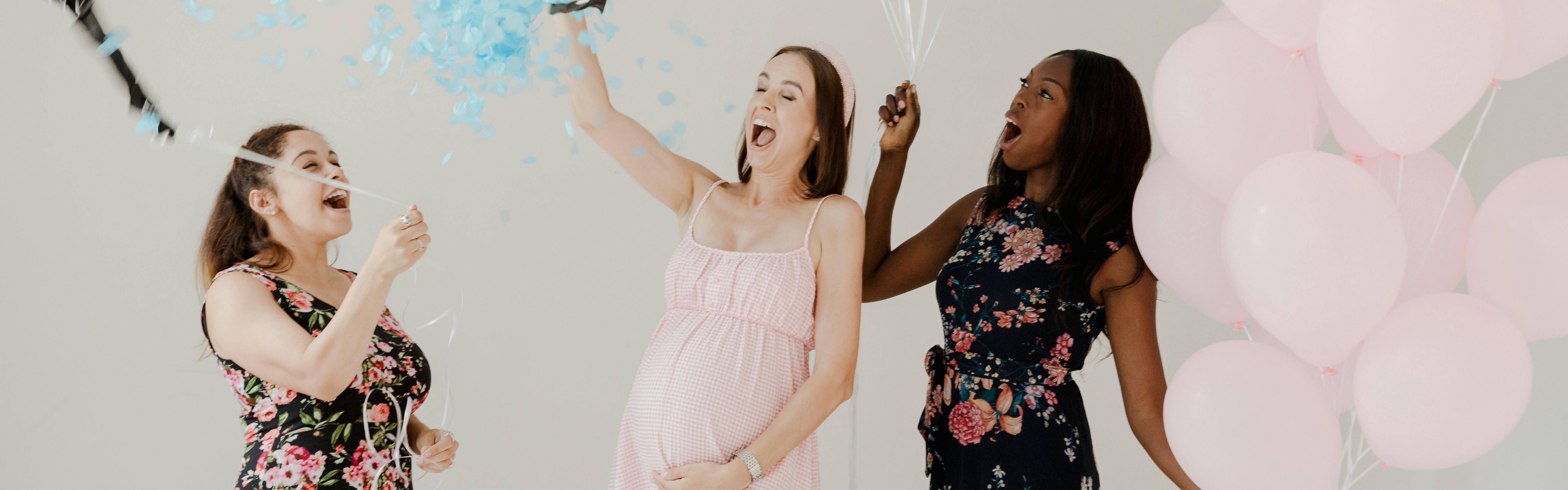 Three women excitedly laughing. There is confetti and one of the woman is holding balloons. One of the women is pregnant. 