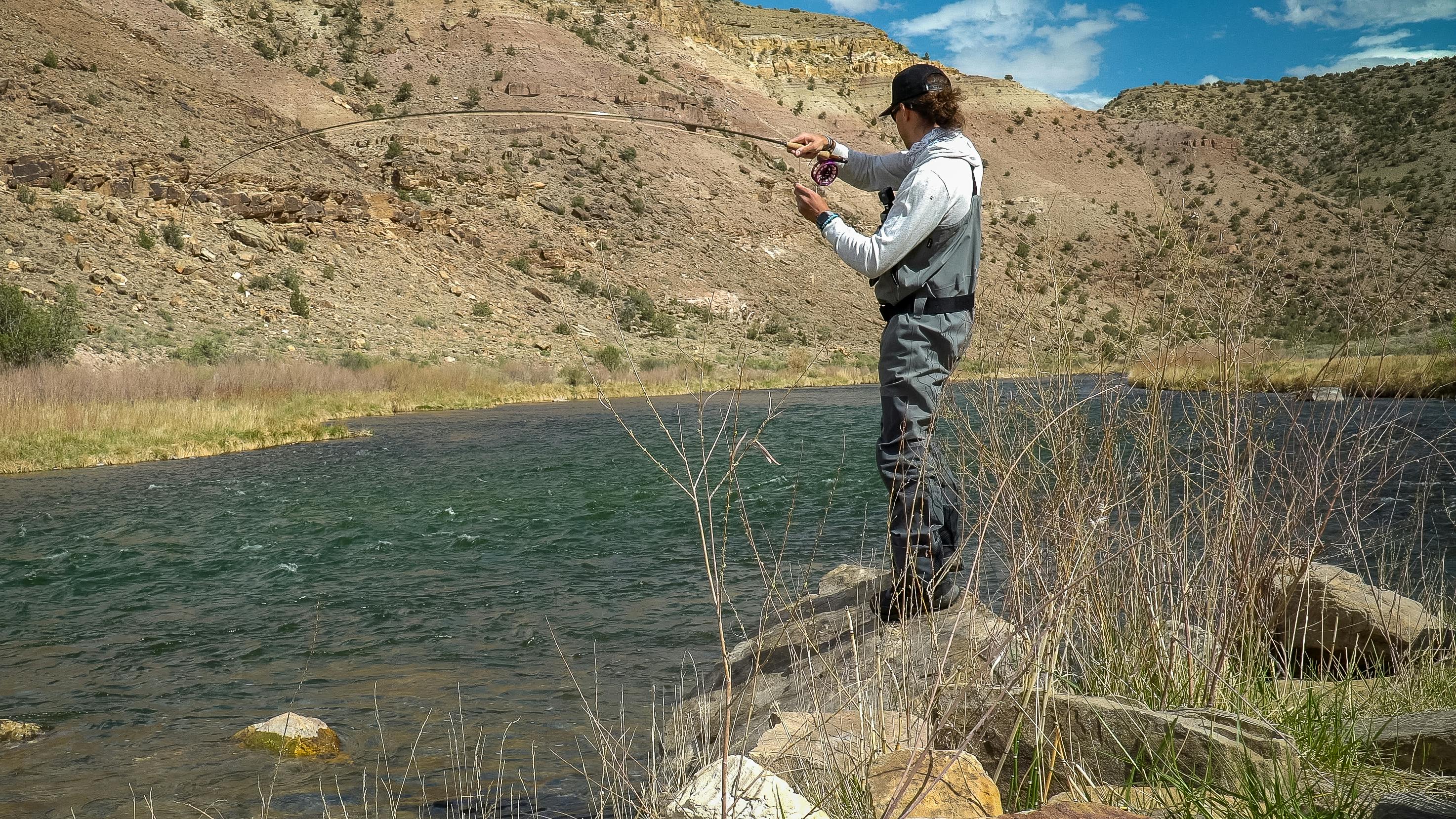 A fisherman standing on a rock and casting a fishing rod into a river.
