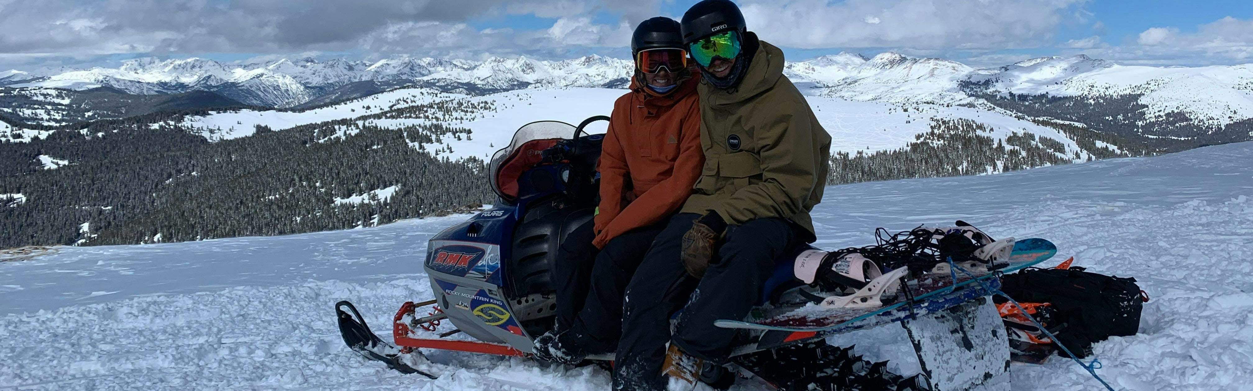 Two people sitting on a snowmobile with ski gear on. There are mountains in the background and one of the people is wearing the Giro Ledge MIPS Helmet.