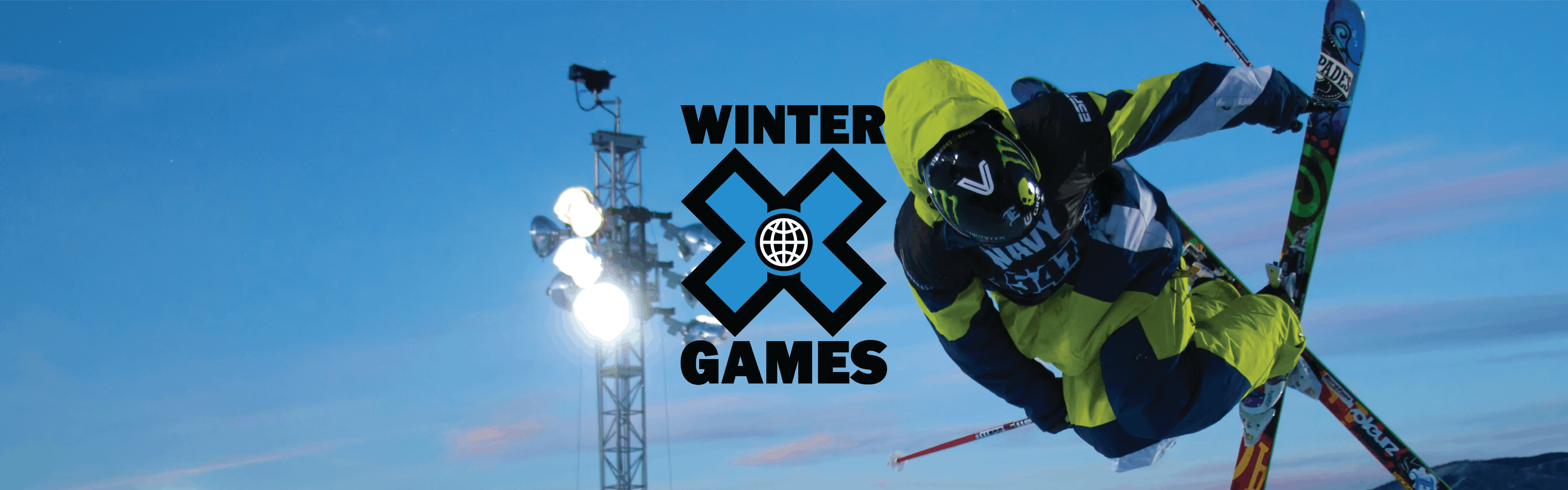 A skier in green jumps and crosses their skis. Behind them is a stadium light. The Winter X Games logo is laid on top.