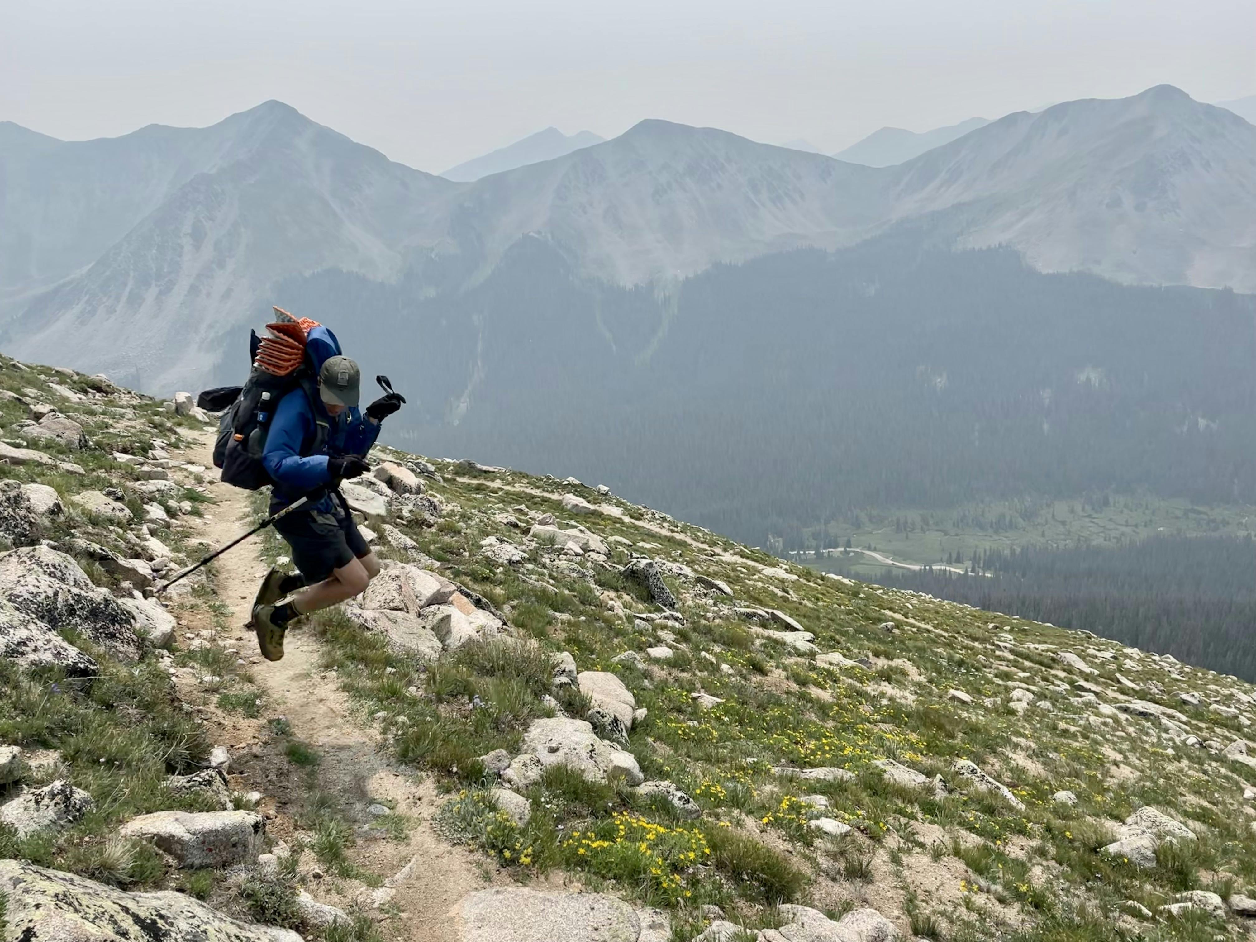 A backpacker with a backpacking setup jumps on the trail. There are mountains in the background.