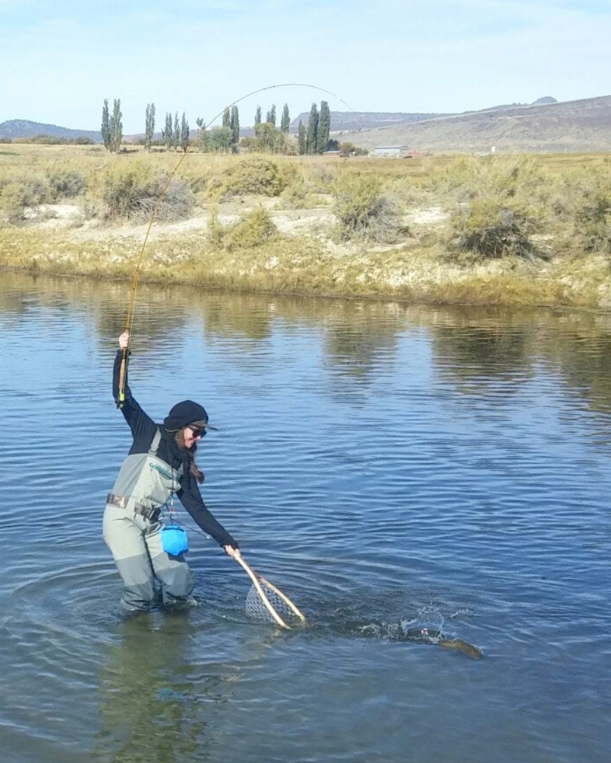 A woman holds a fly fishing rod in one hand and a net in the other while trying to catch a fish