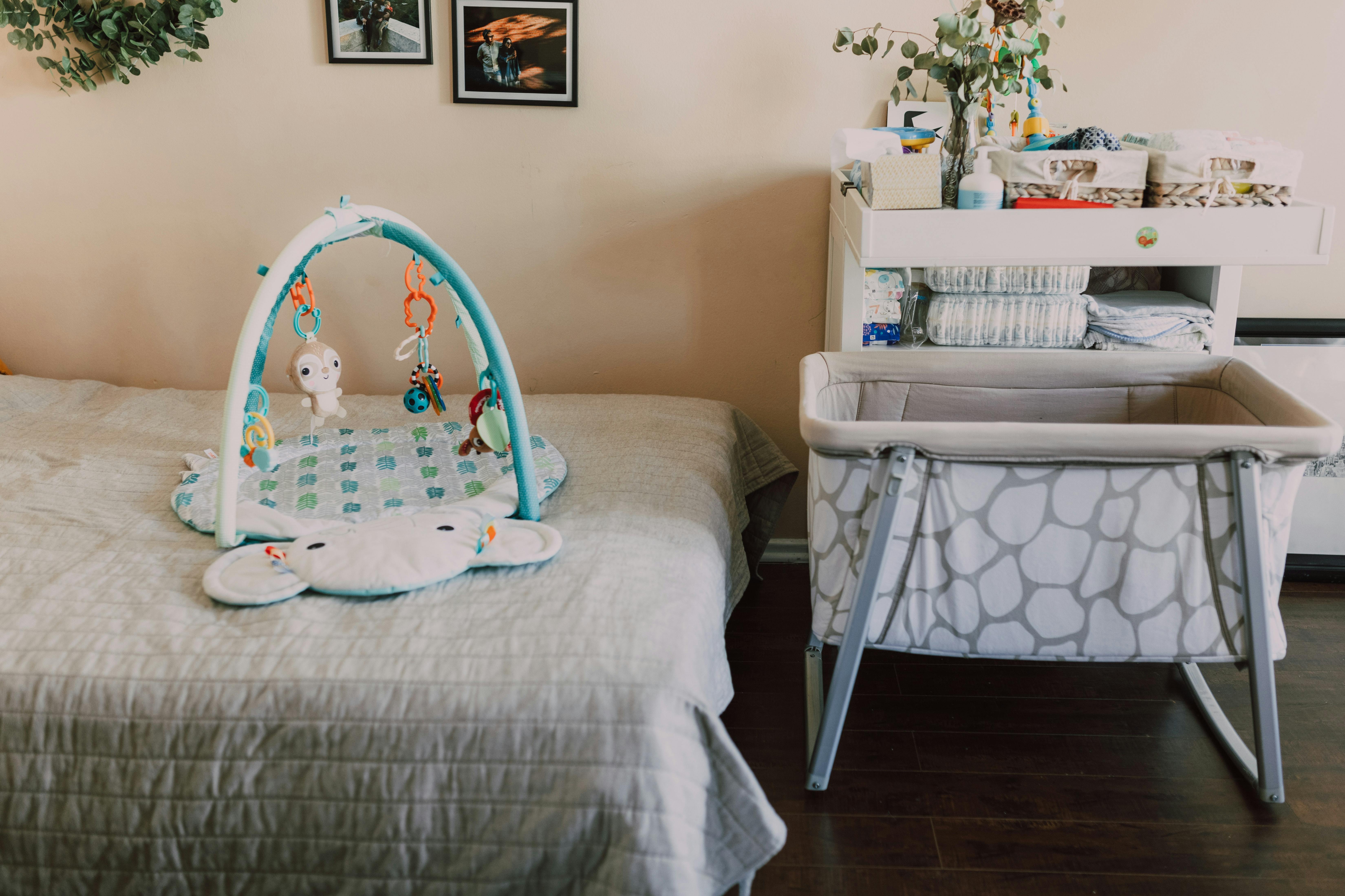 A travel crib is setup next to a bed. On the bed is an activity center for a baby. 