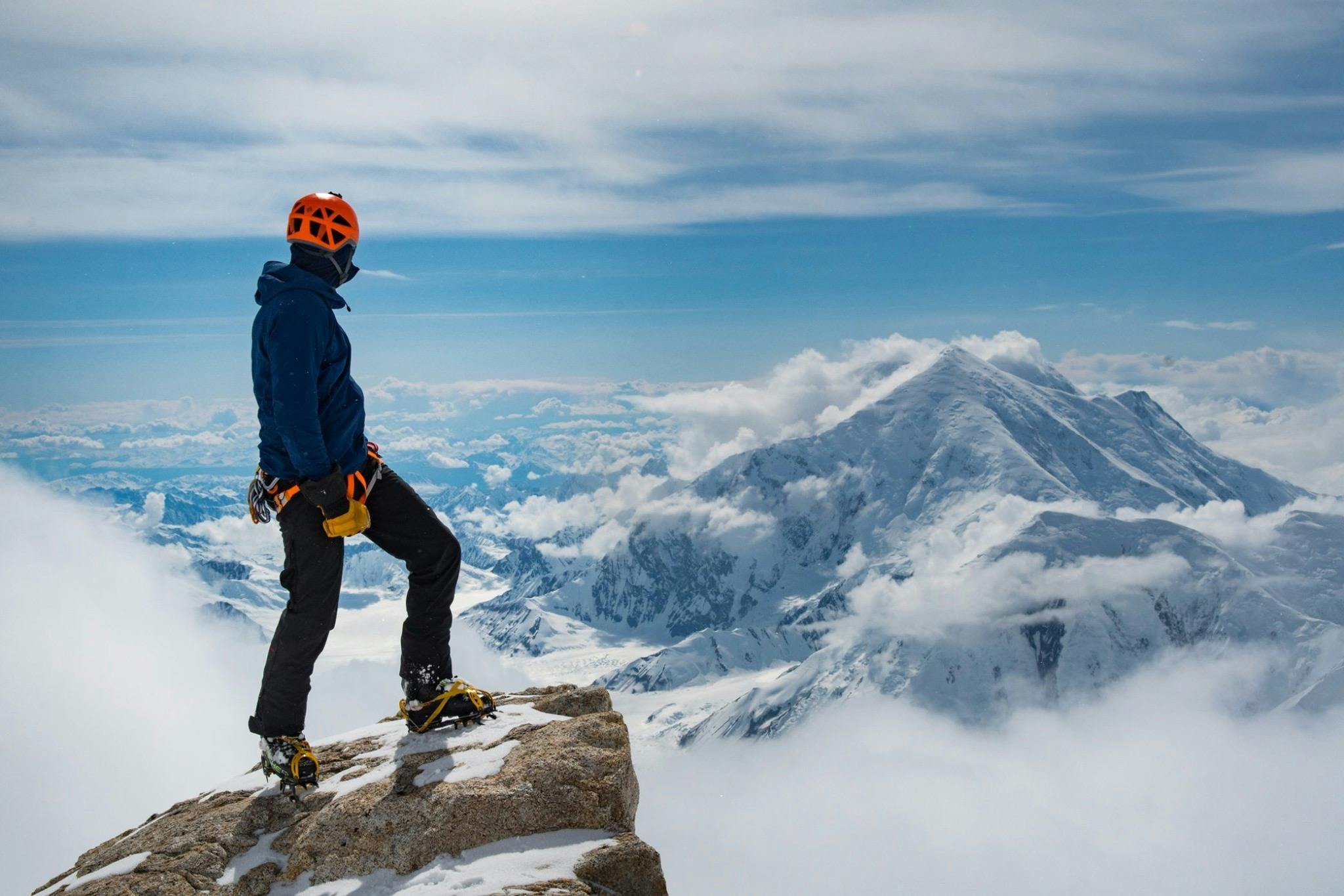 A skier wearing crampons and a harness stands at the top of a mountain. There are mountains in the background. 