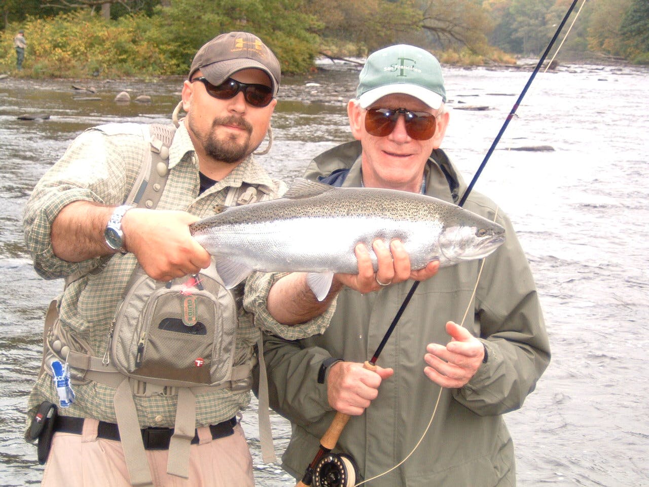 Two fishermen stand in the water. One holds a fly rod and the other holds a steelhead. 