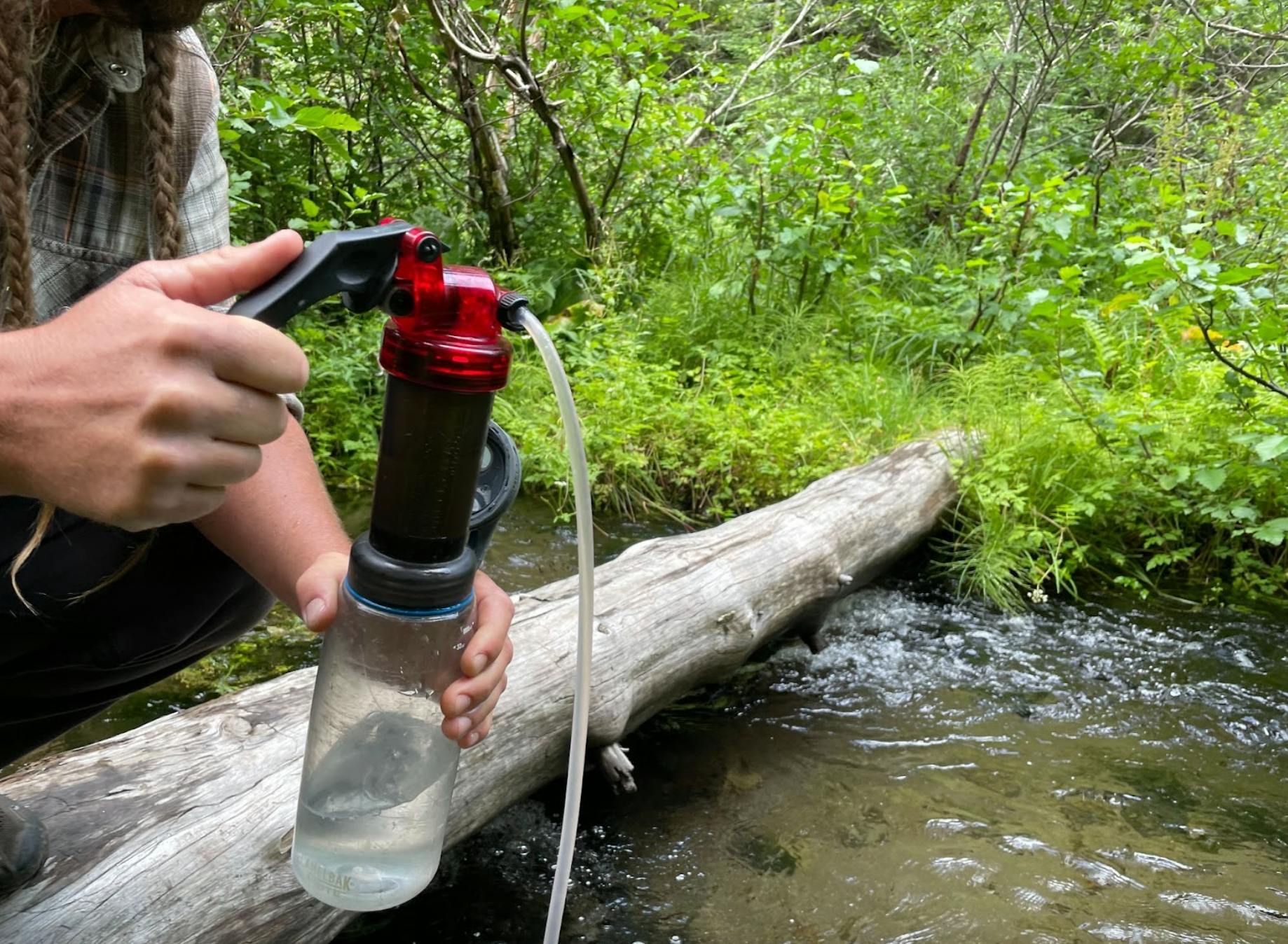 The author's partner pumps water into their water bottle with the MSR Miniworks EX filter as they squat on a log crossing a creek.