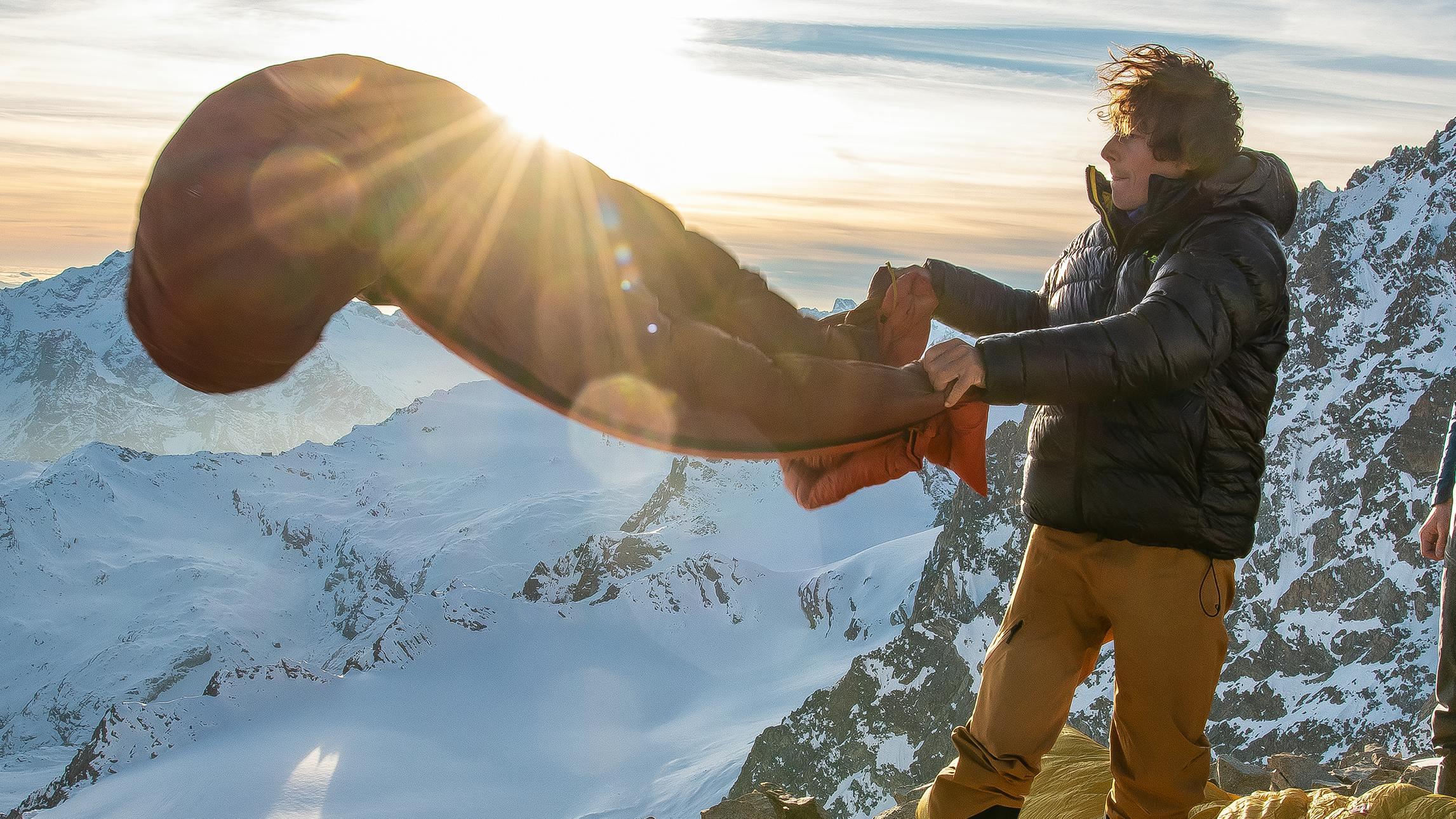 A hiker fluffy out his sleeping bag on a mountain summit next to his hiking partner with the sun setting over snowy mountain scenery in the background.