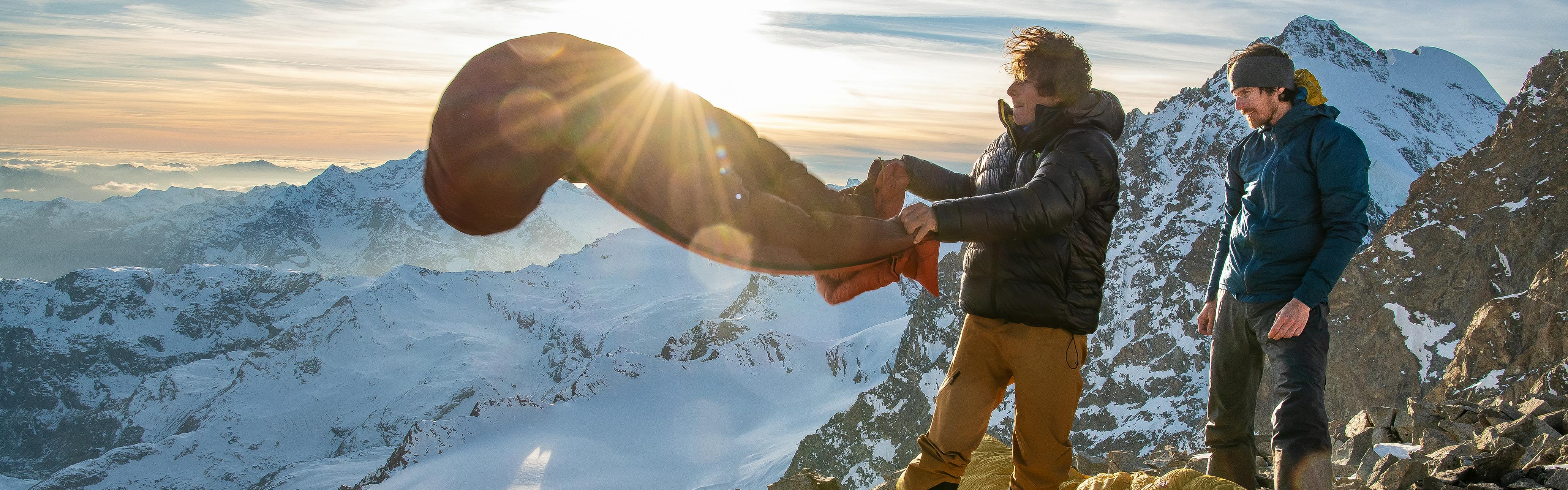 A hiker fluffy out his sleeping bag on a mountain summit next to his hiking partner with the sun setting over snowy mountain scenery in the background