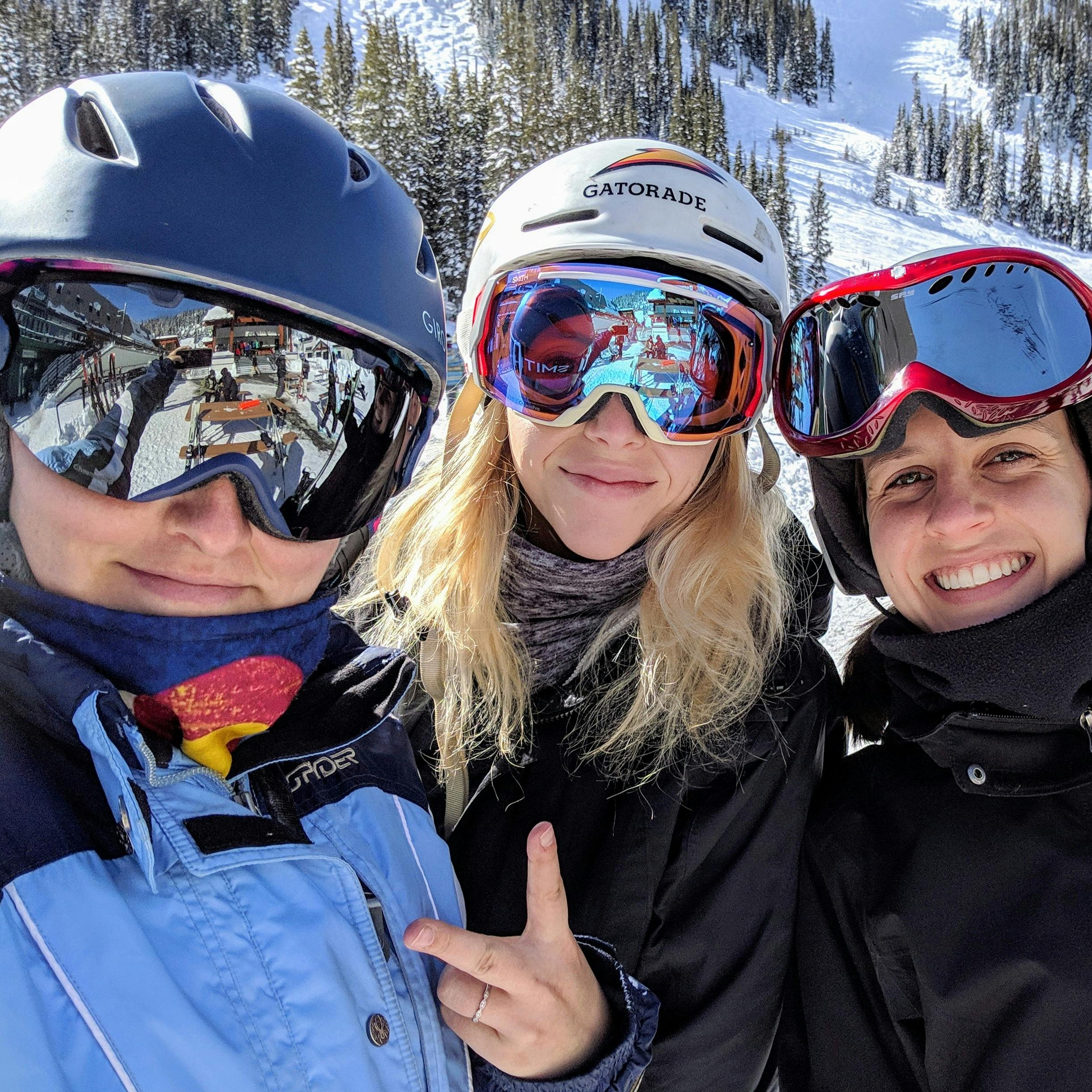 Three skiers wearing helmets are all smiling at the camera. There is a ski run visible in the background. 