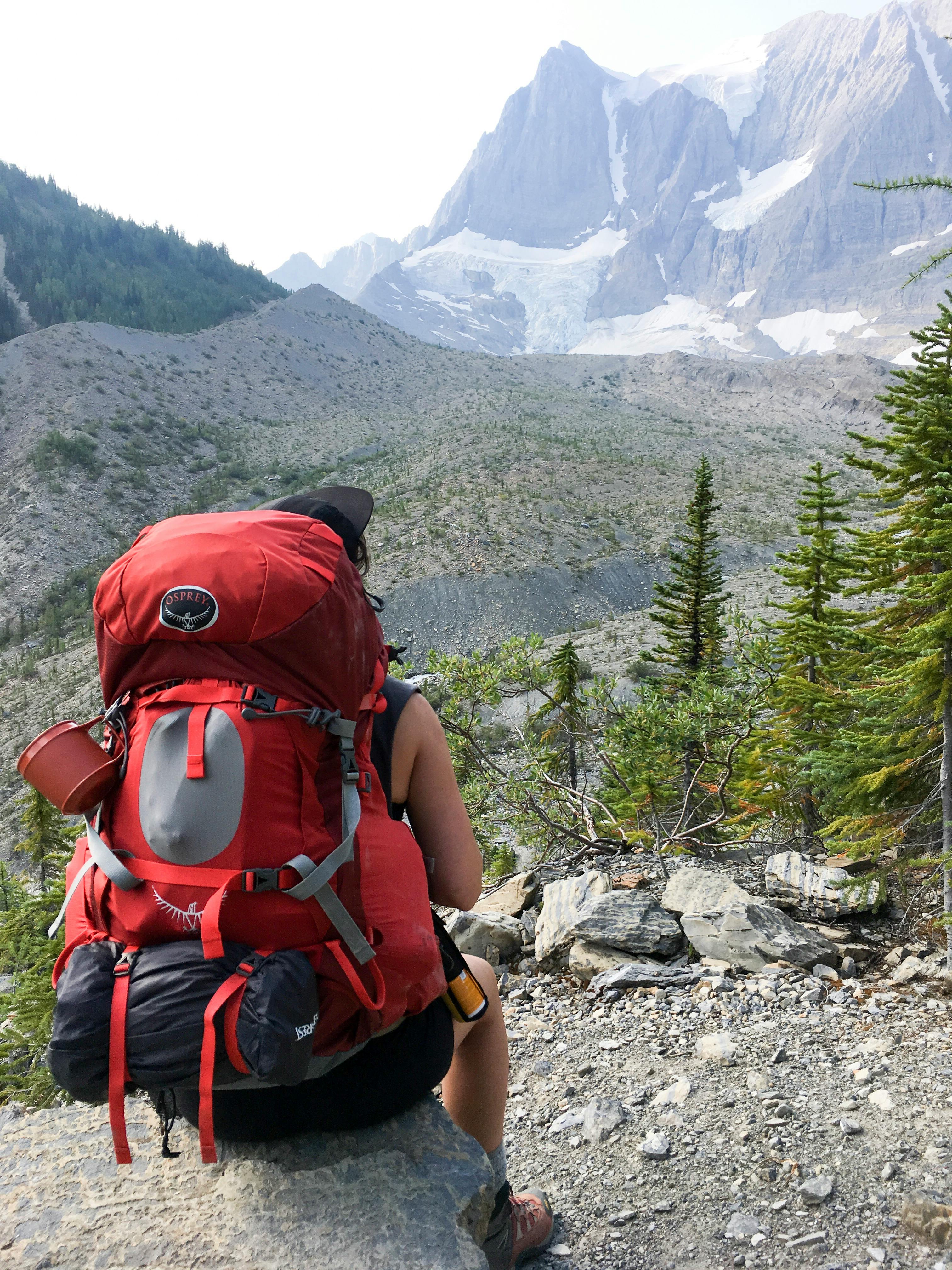 A person wearing a bright red backpacking pack sits facing away from the camera and towards mountains and trees