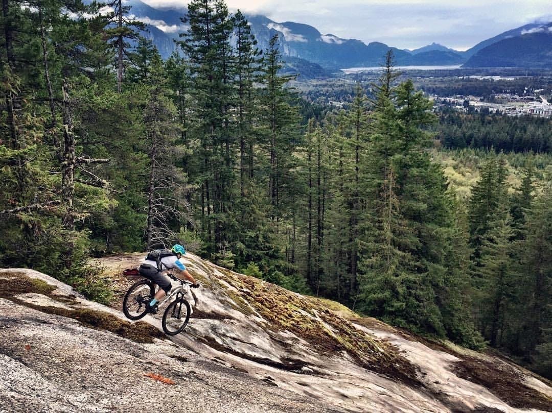 A man mountain biking down a granite slope in Squamish, Canada with mountains and a lake in the distance.