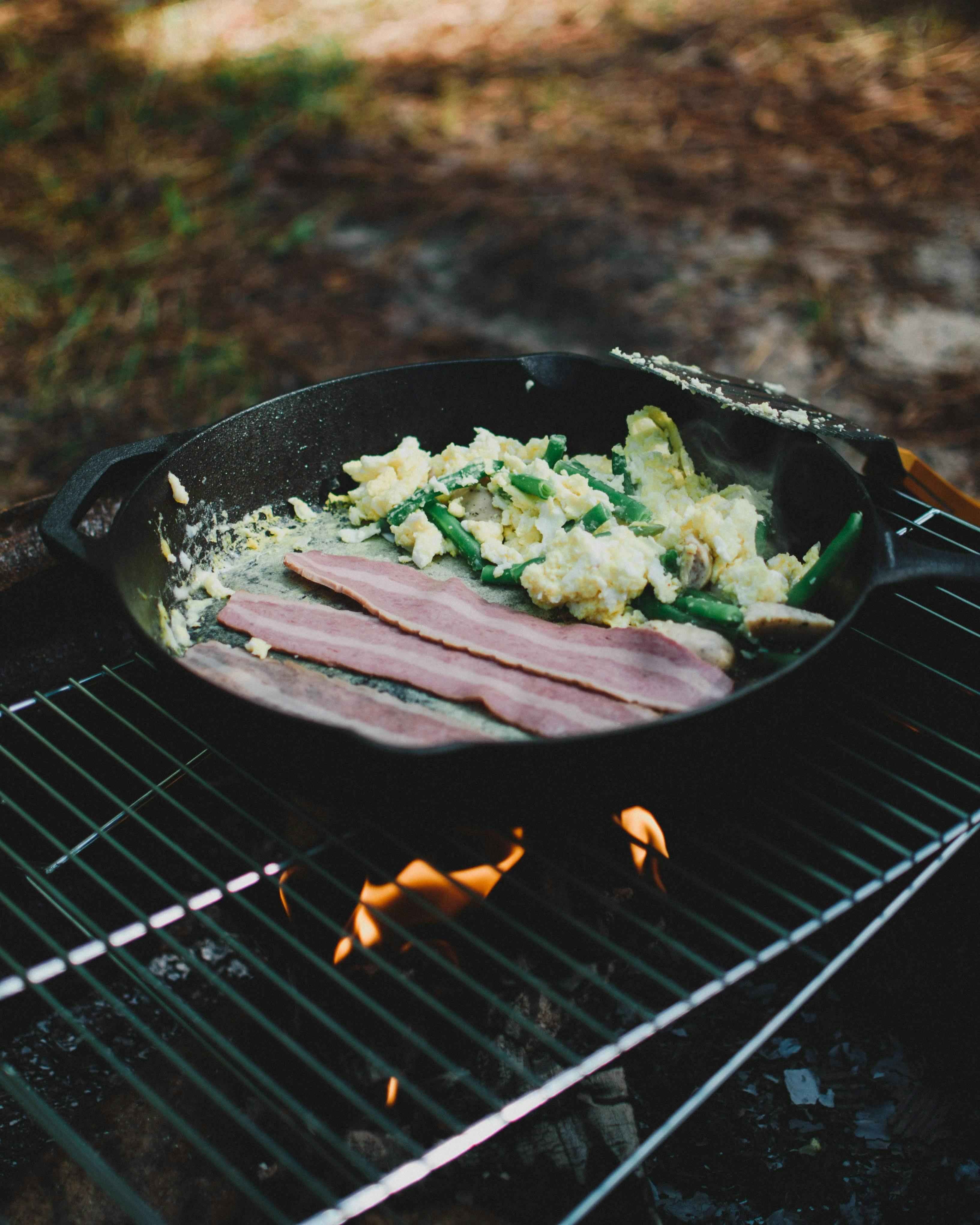 A cast-iron skillet sits on a grate above a campfire. In the skillet is a scramble with a few slices of bacon.