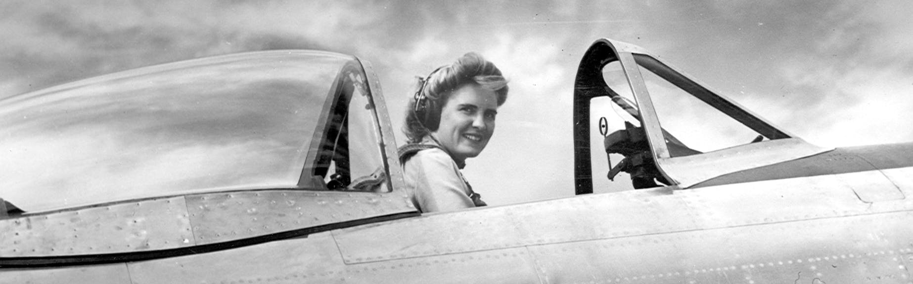 Celia Hunter sits in the cockpit of an open plane and smiles. The image is in black and white.