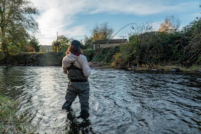 A fisherman standing in a river with waders. They have a fish on their line and the rod is bending. 