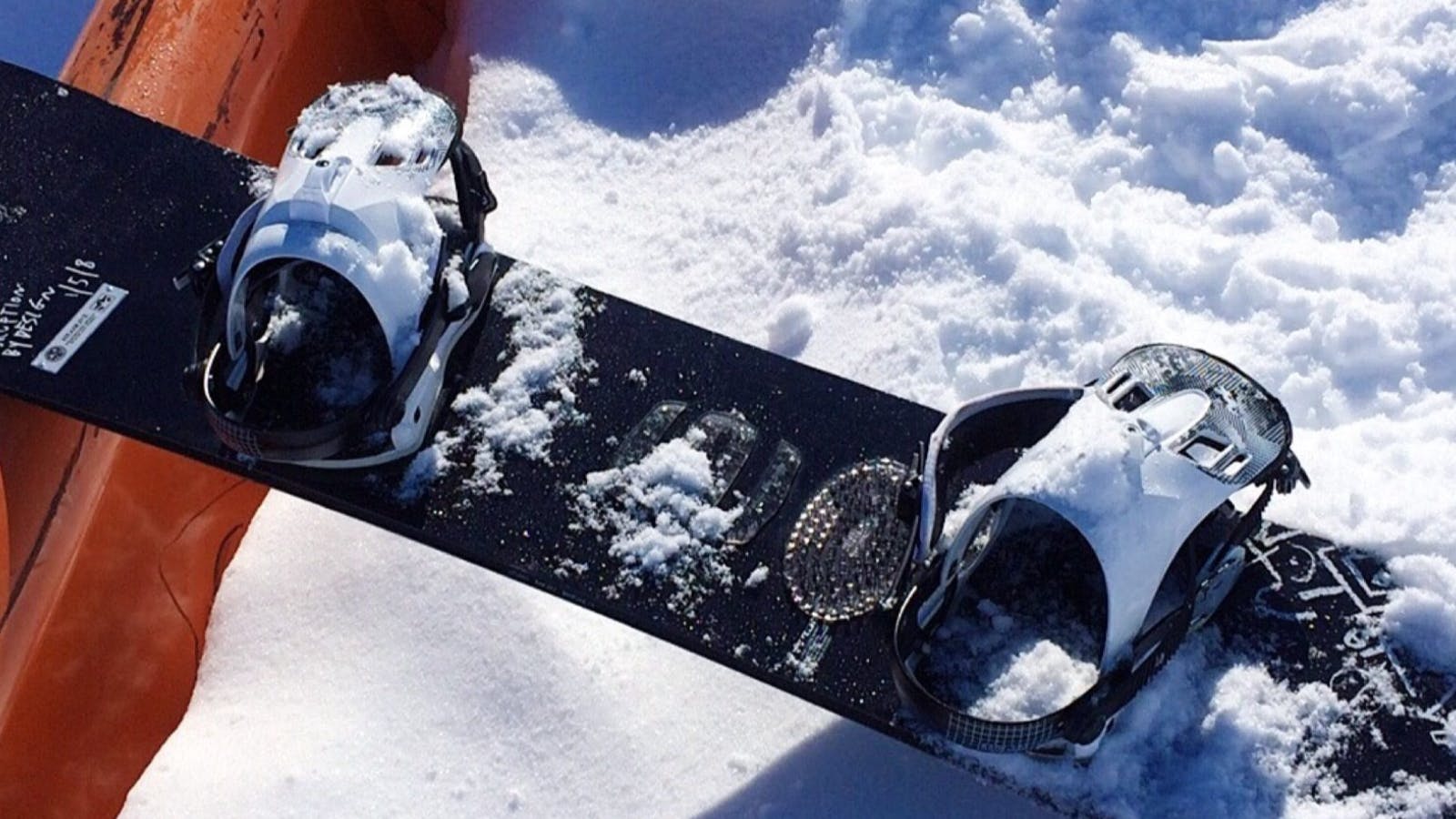 A snowboard with bindings sits in the snow, propped against a rail. 