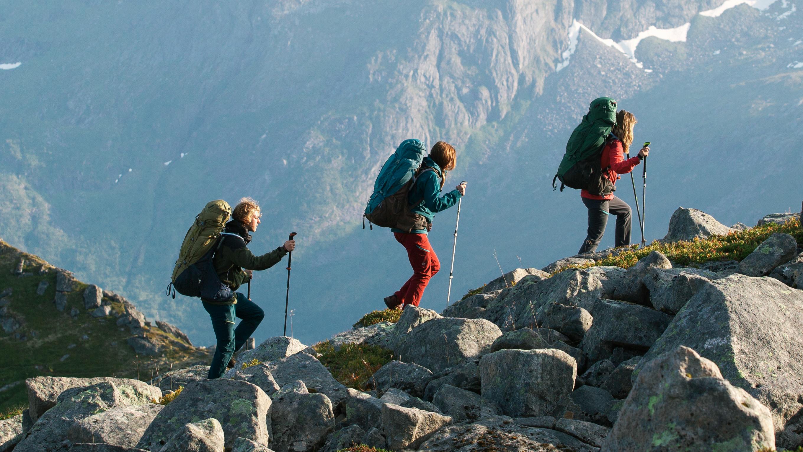 Three people backpack up a hill covered in boulders. They all wear large Deuter backpacks and carry hiking poles.