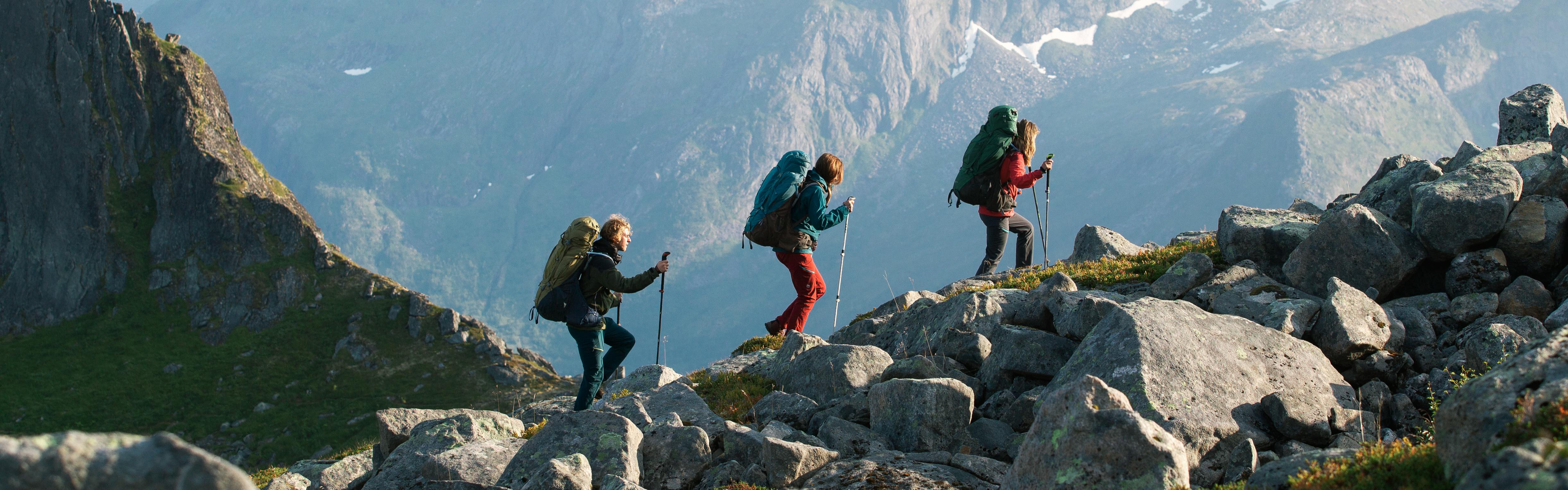Three people backpack up a hill covered in boulders. They all wear large Deuter backpacks and carry hiking poles.