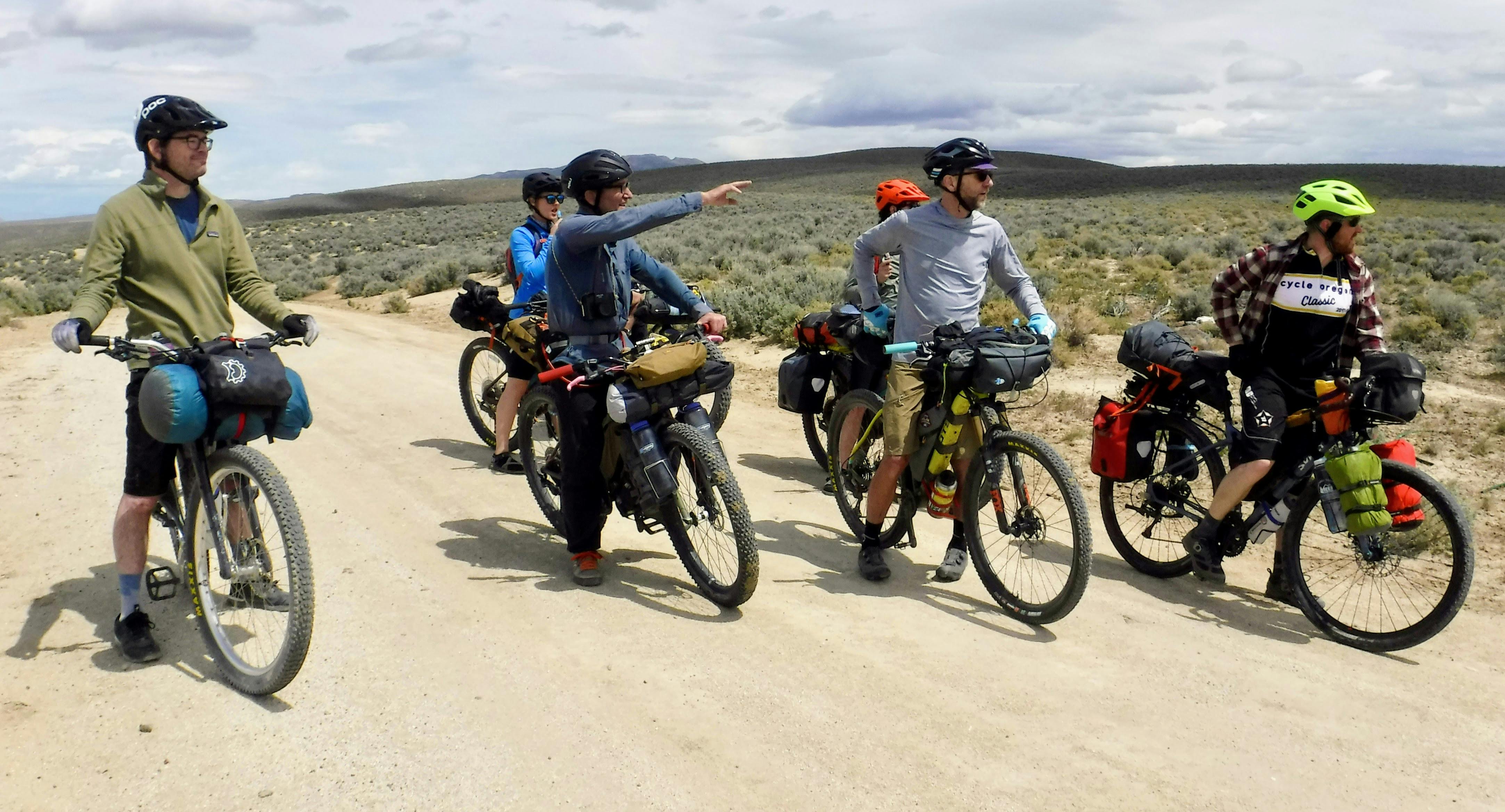Six bikepackers stand on a dirt trail. One is pointing down the trail. 