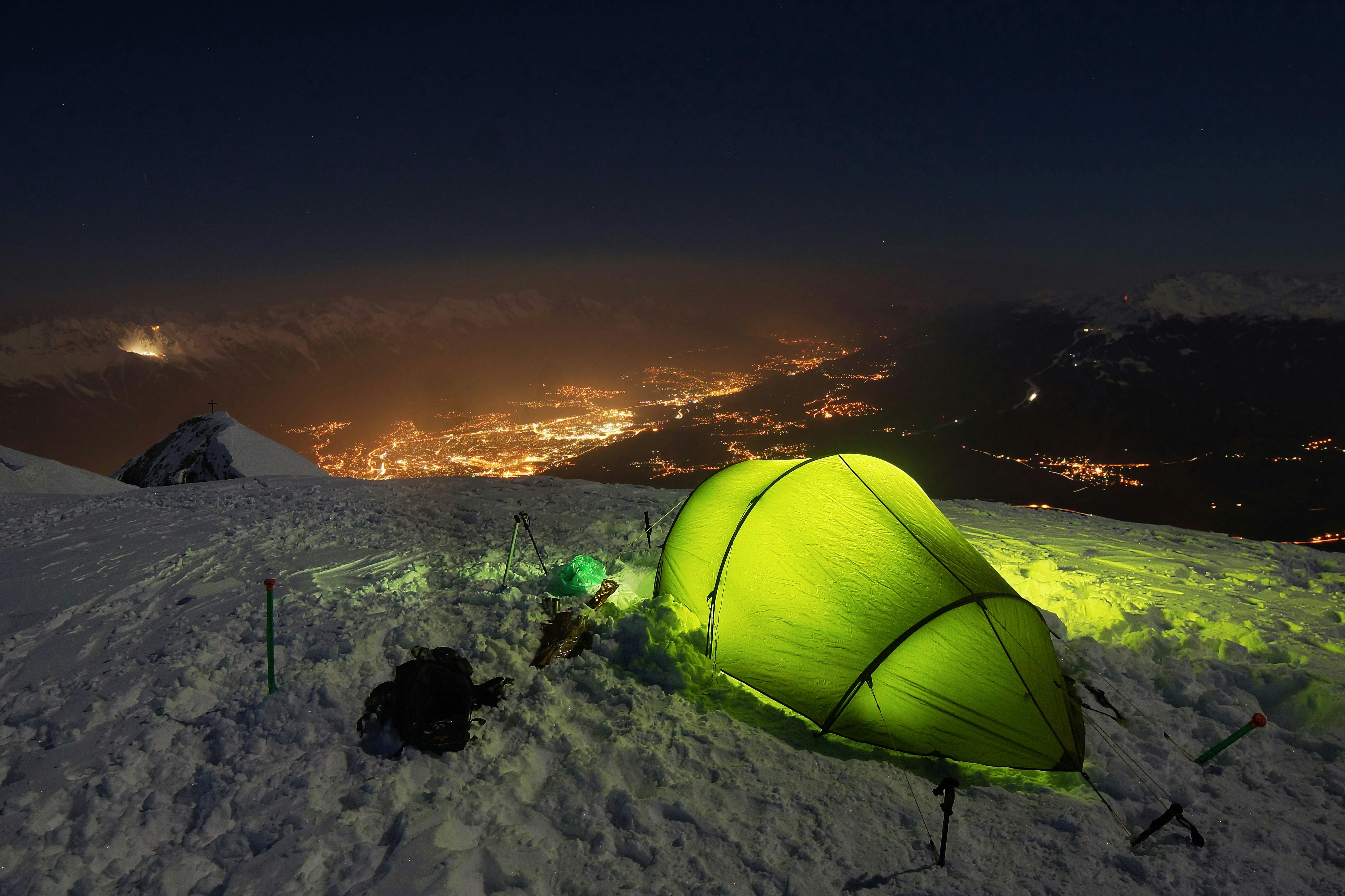 A light illuminating a bright green tent from within. The tent is set up on the snow, the night sky and brightly lit city in the valley in the distance