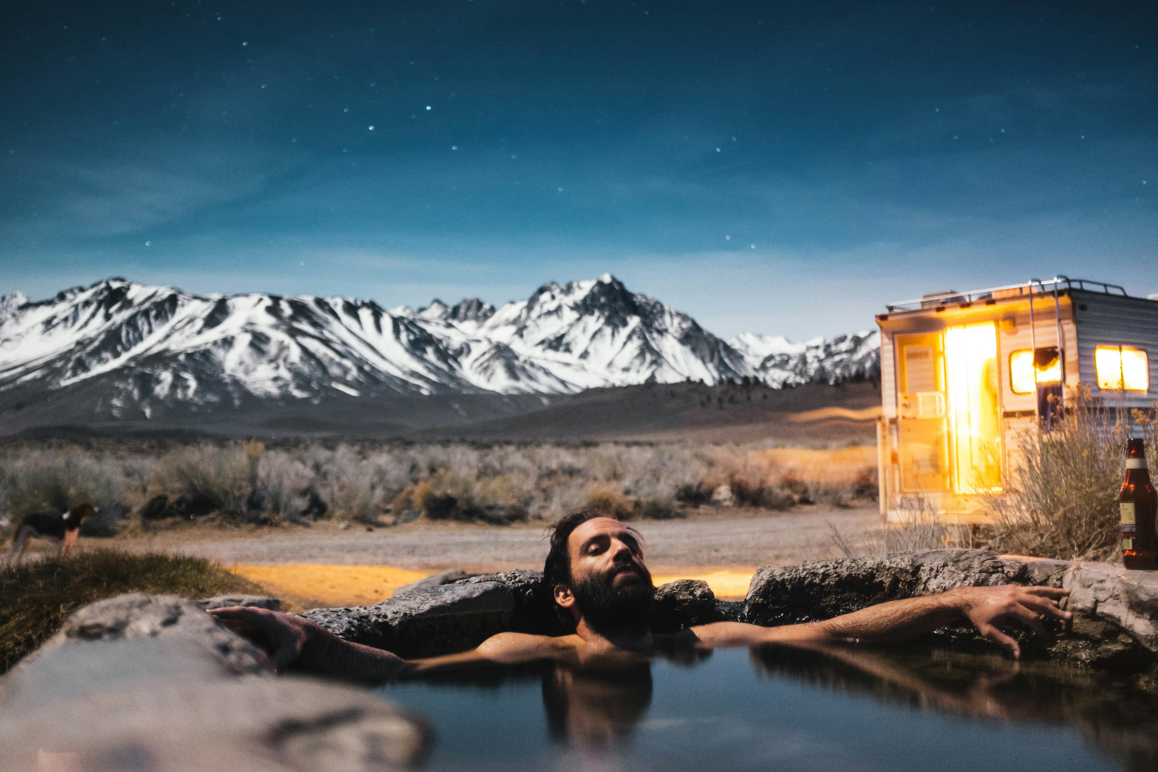 Man relaxing in a natural hot spring with his campervan and snowy mountains in the background