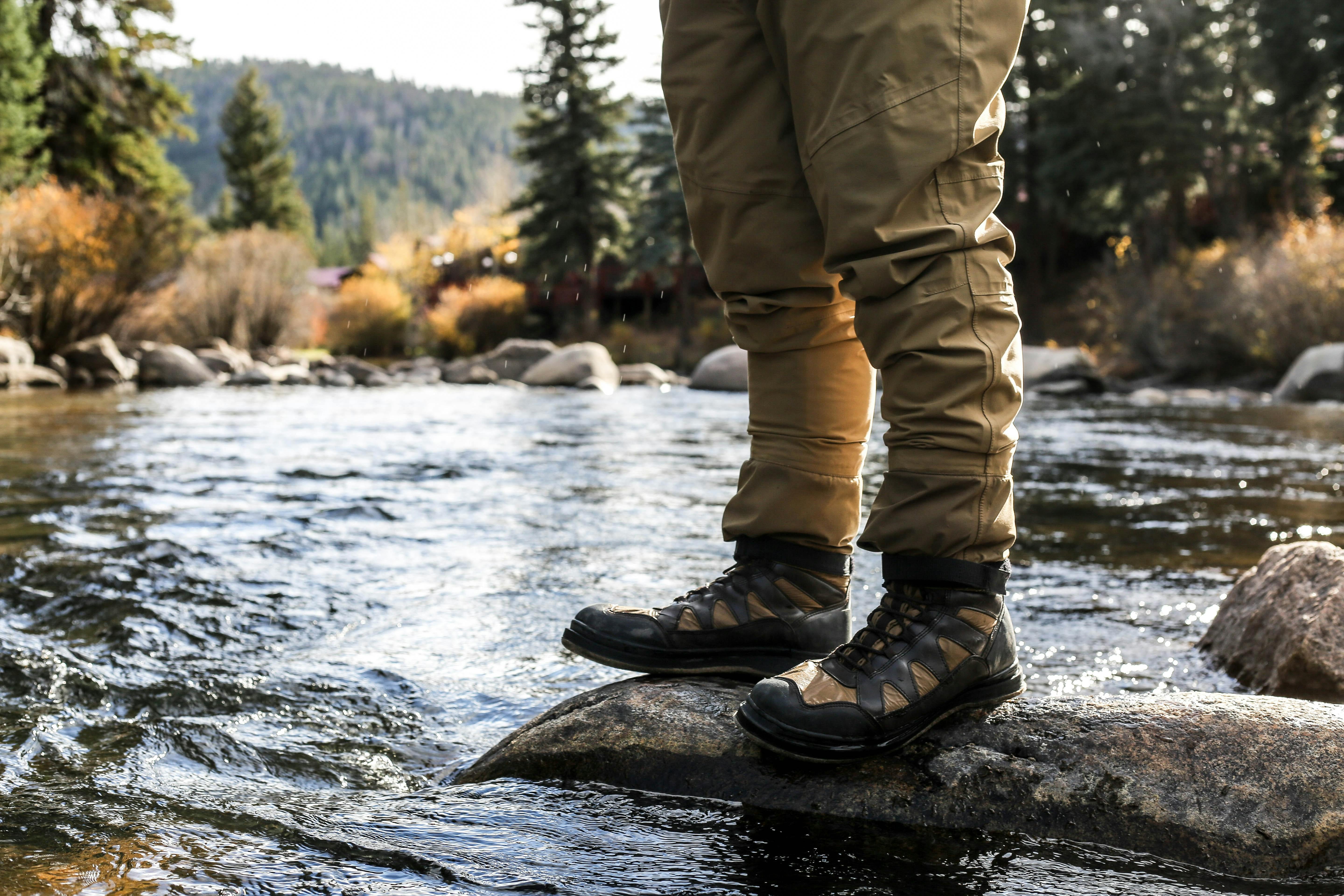 A man with fishing boots and waders stands at the edge of a river.