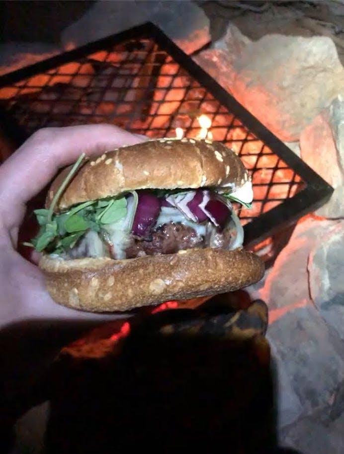 Someone holds a burger over a grill on top of a campfire. The burger has melted cheese, onions, and arugula on it. 