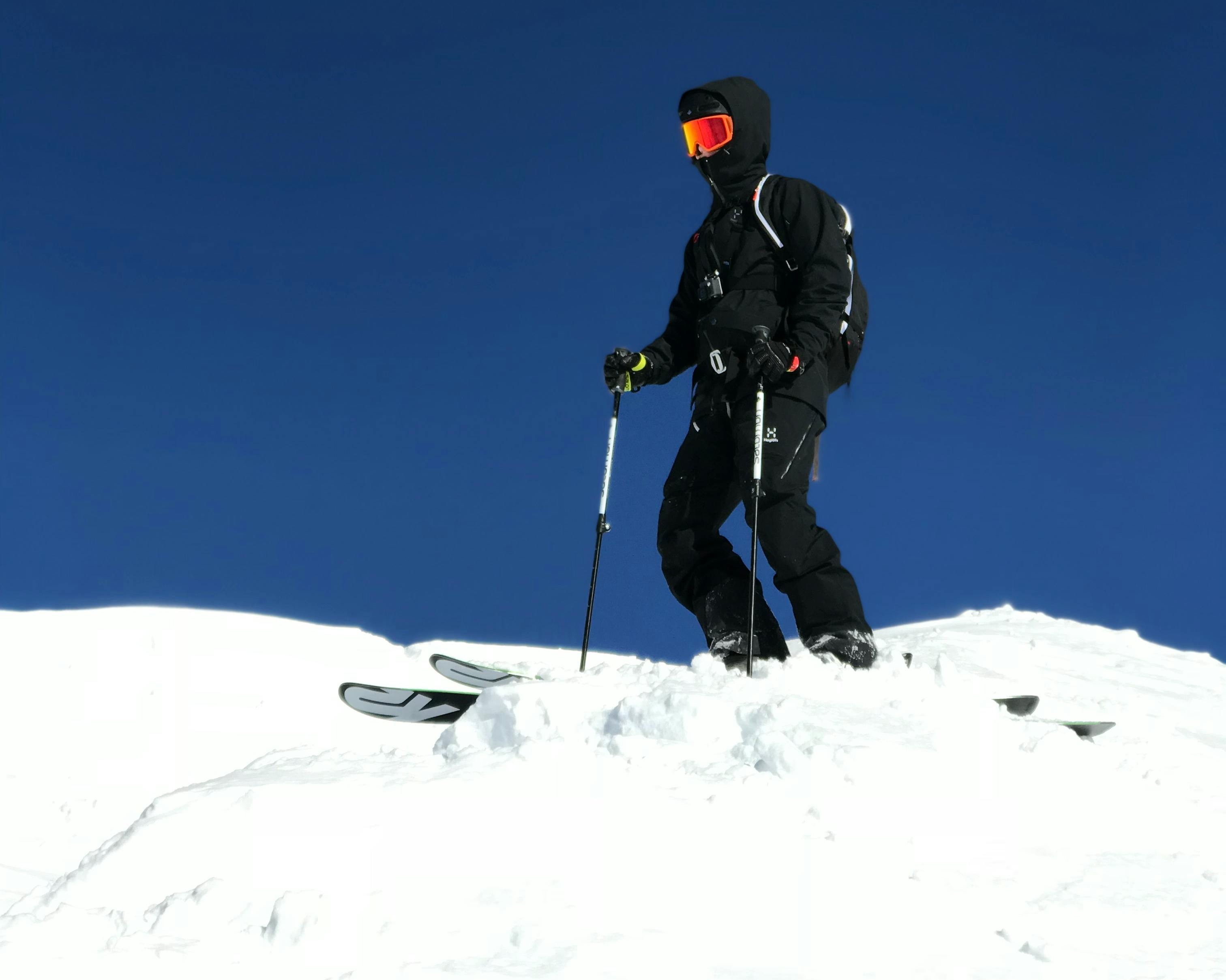 A skier in a black jacket and black ski pants at the top of a ski slope under a clear blue sky