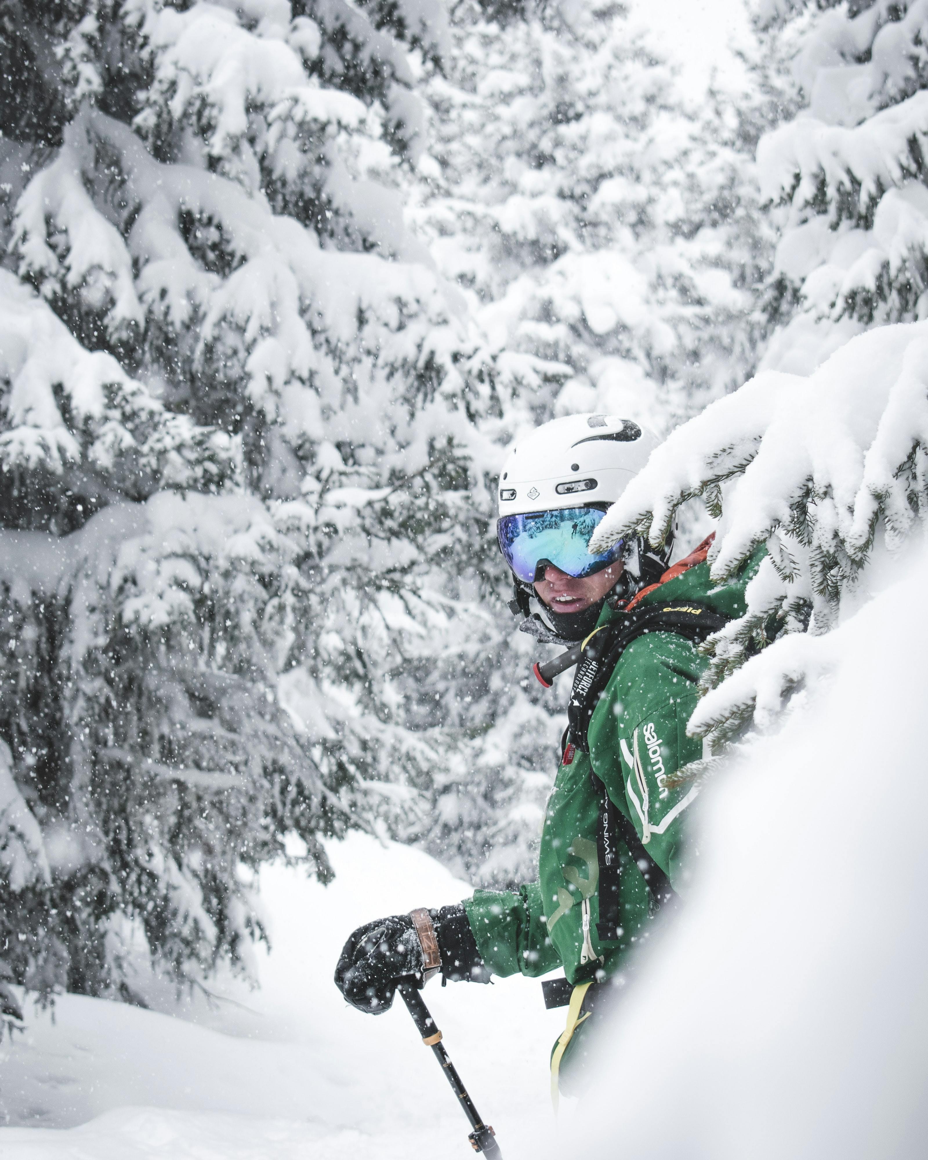 A skier standing in between some snowy trees. He is wearing a helmet and green ski jacket. 