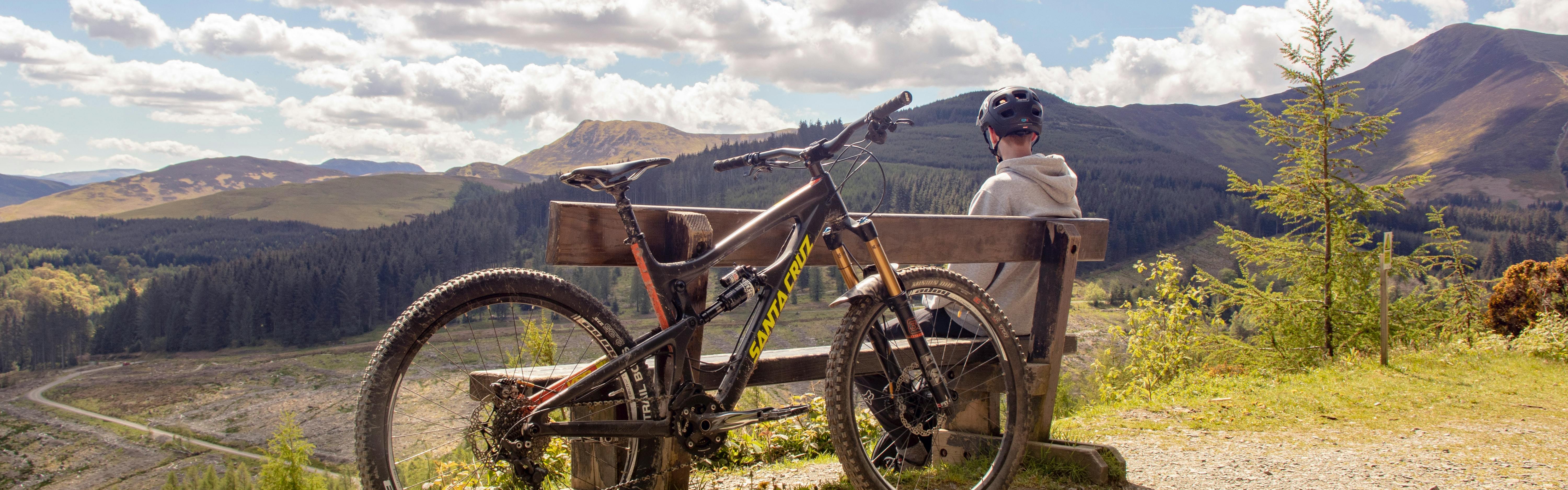 A biker sits on a bench on a mountain while his bike rests on the bench behind him. 