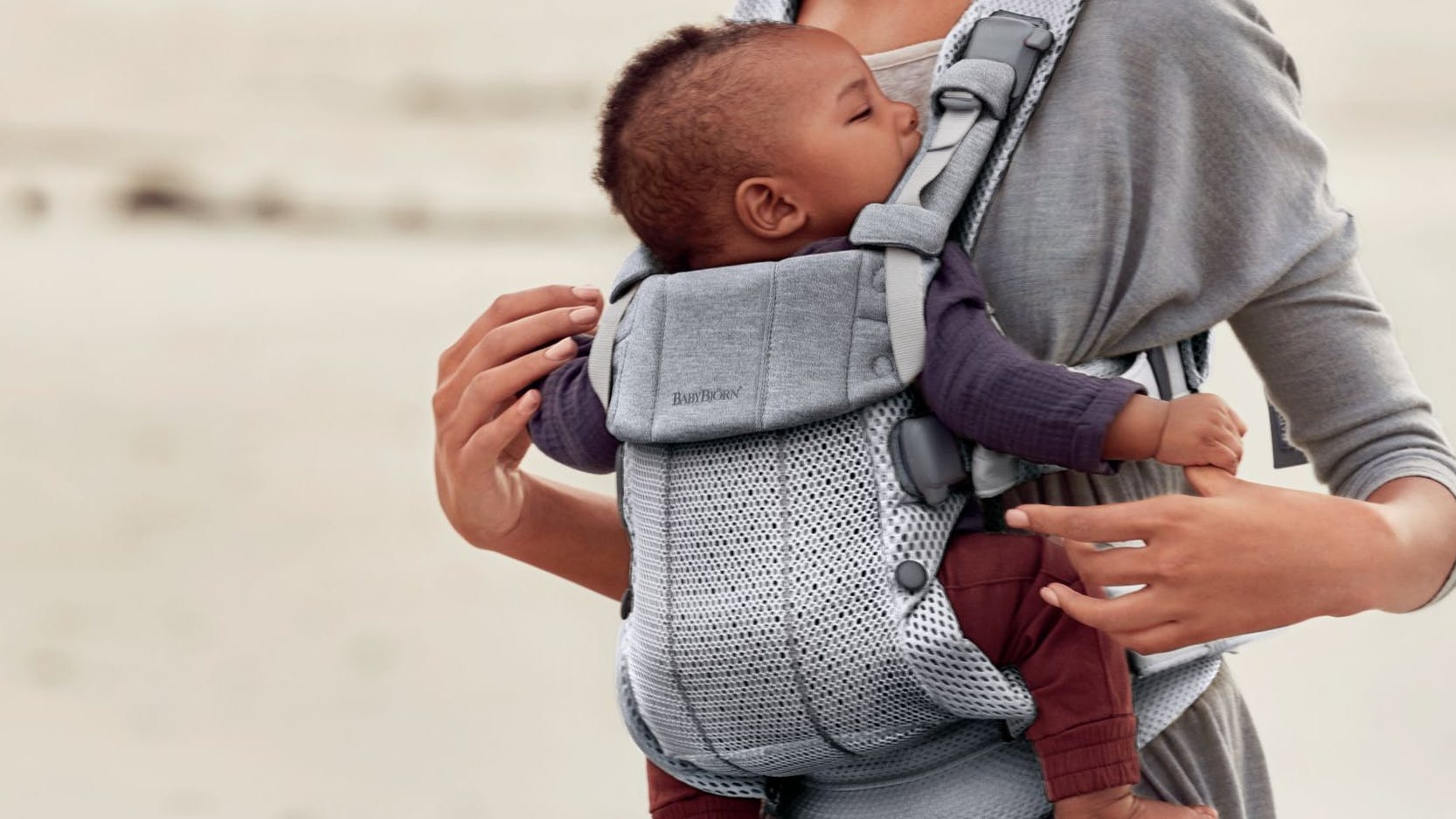 A woman walks on the beach while holding a baby in the BabyBjörn Baby Carrier Harmony.
