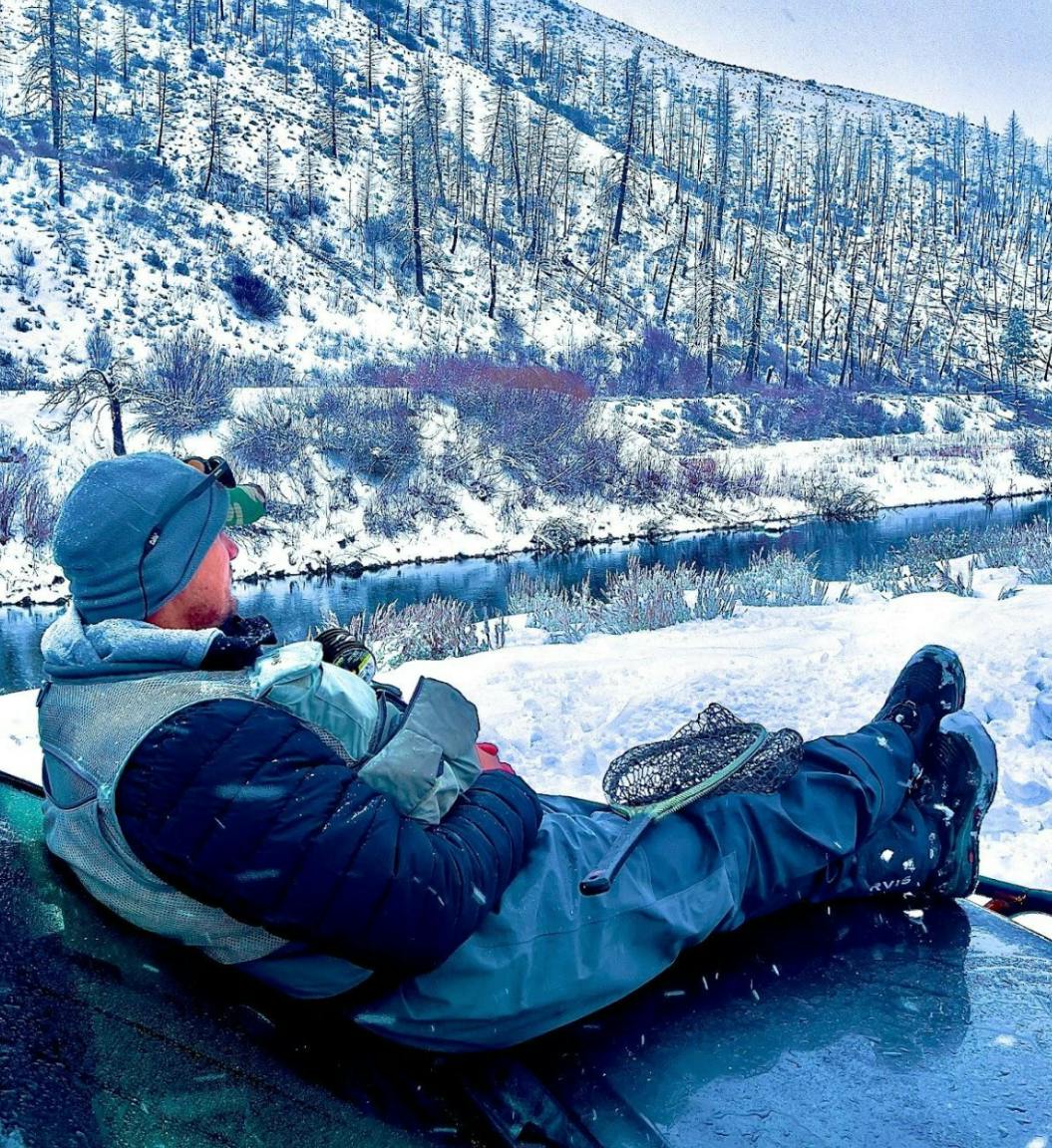 A fisherman sitting on his car while looking out at a snowy river.