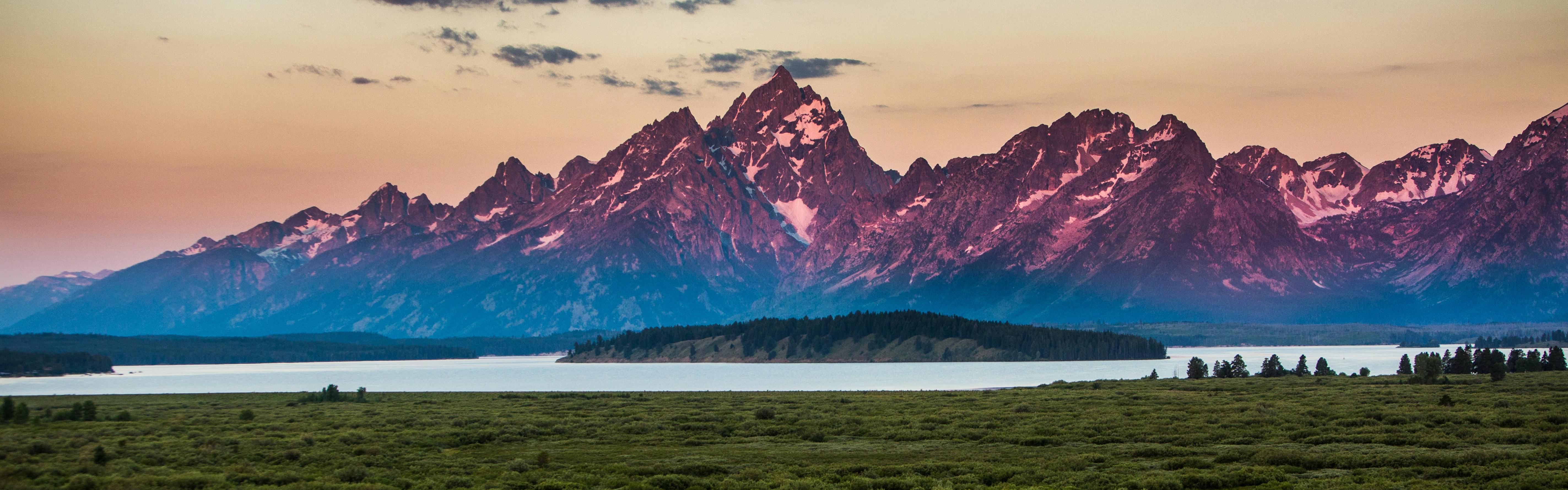 The view from Jackson Lodge, Grand Tetons National Park. There's an open meadow, a lake with an island beyond it, and the Grand Tetons in the distance, purple in the sunrise. 