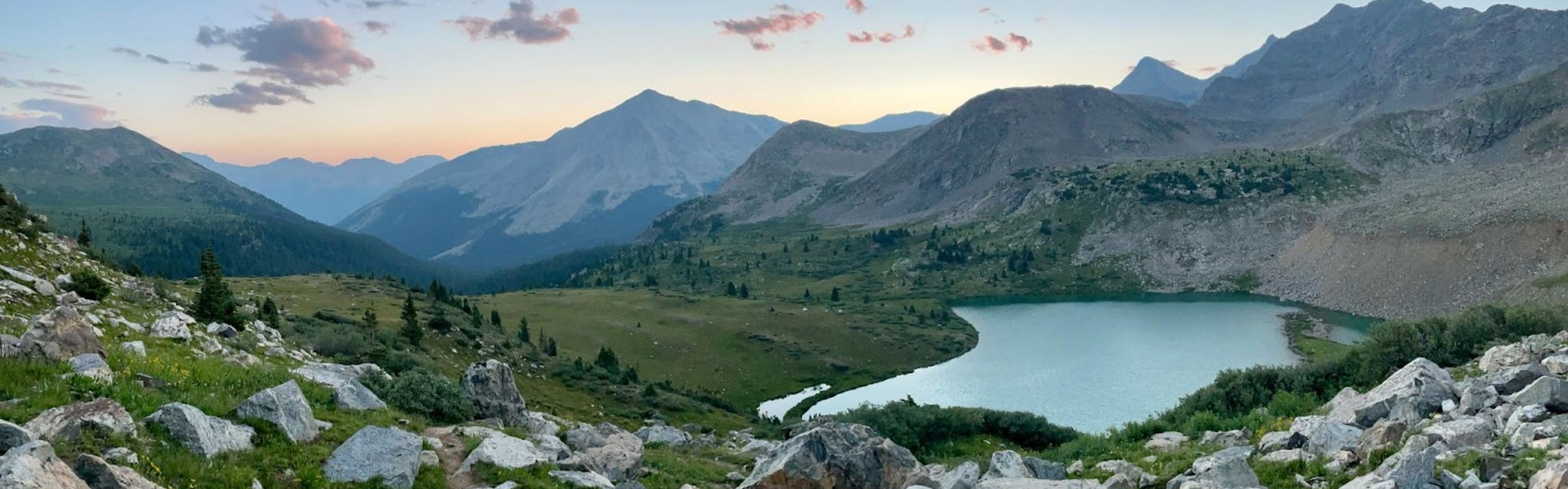 A high alpine lake with mountains, trees, and boulders. There is a sunset happening in the background.