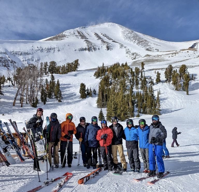 A group of skiers stand at the bottom of a ski run. There is a lot of snow in the background. 