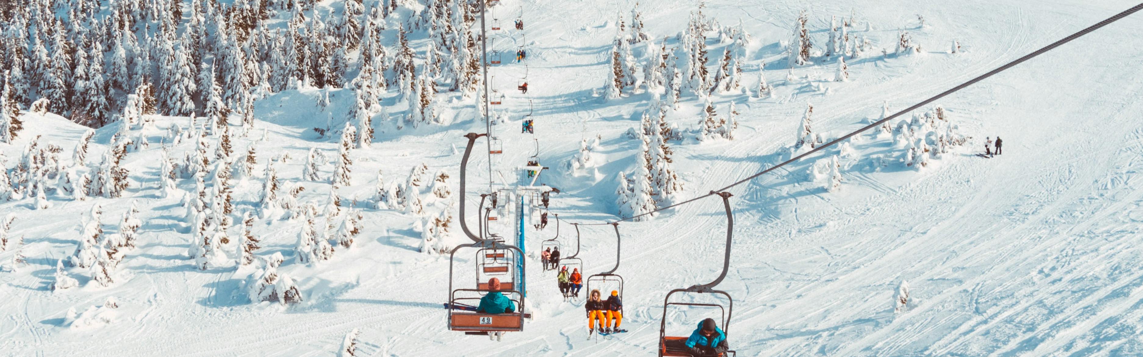 A ski lift with people riding up the mountain. You can see almost the whole lift and it looks pretty busy because there are people in every chair. There is a lot of snow on the ground.