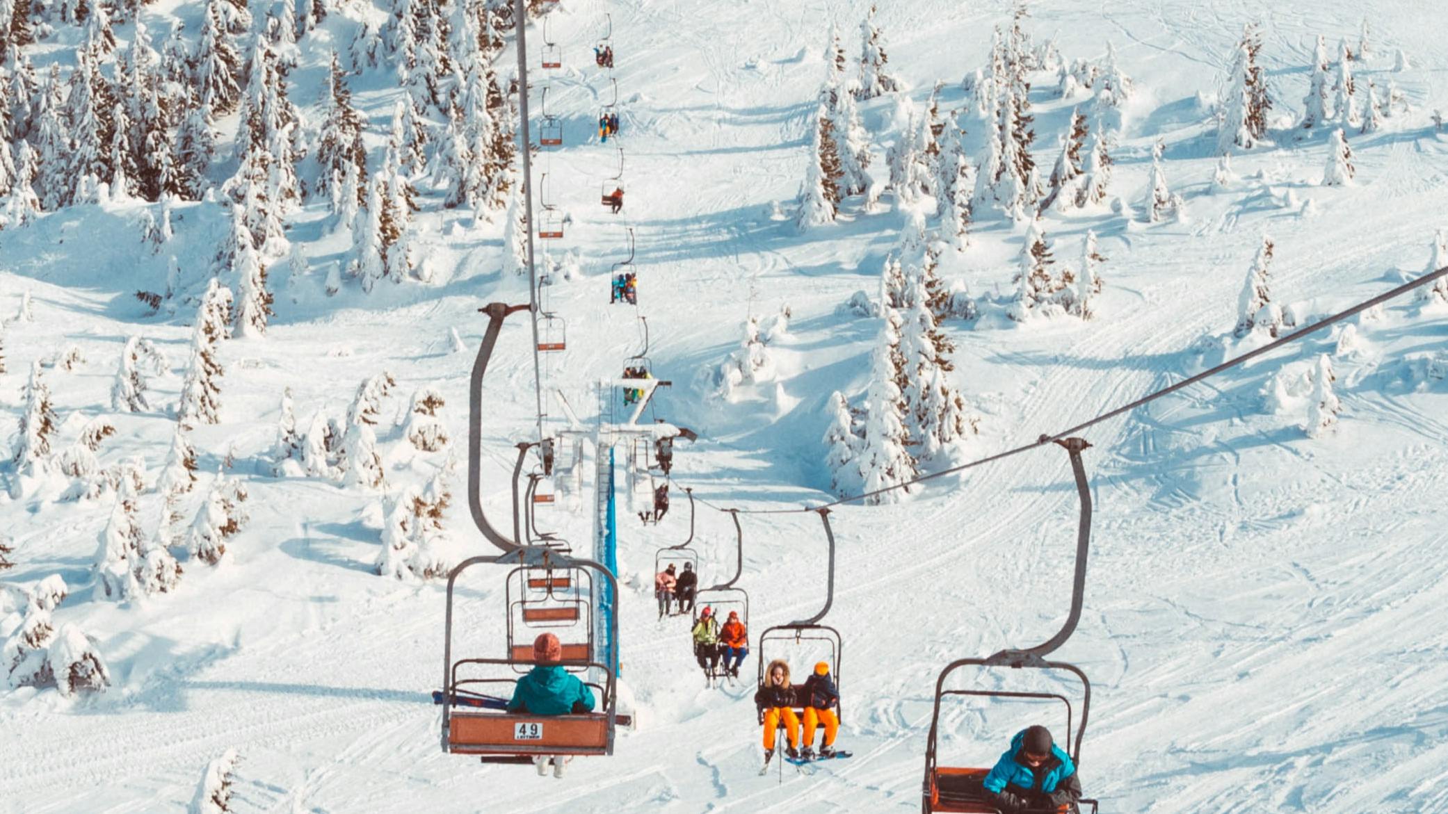 A ski lift with people riding up the mountain. You can see almost the whole lift and it looks pretty busy because there are people in every chair. There is a lot of snow on the ground.