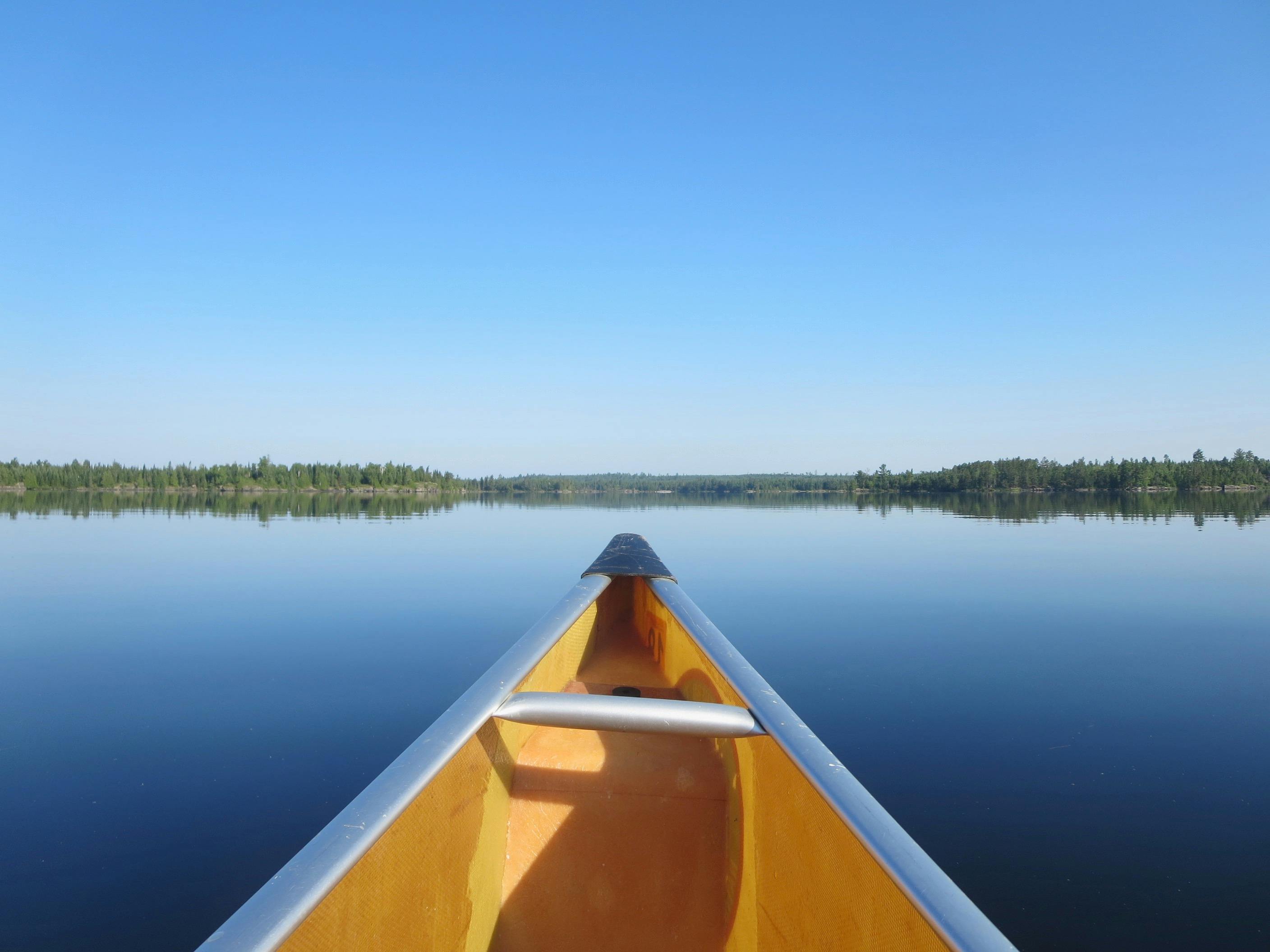 An image of the tip of a yellow canoe on extremely blue, flat, still water with low-lying green foliage on the banks in the distance. 